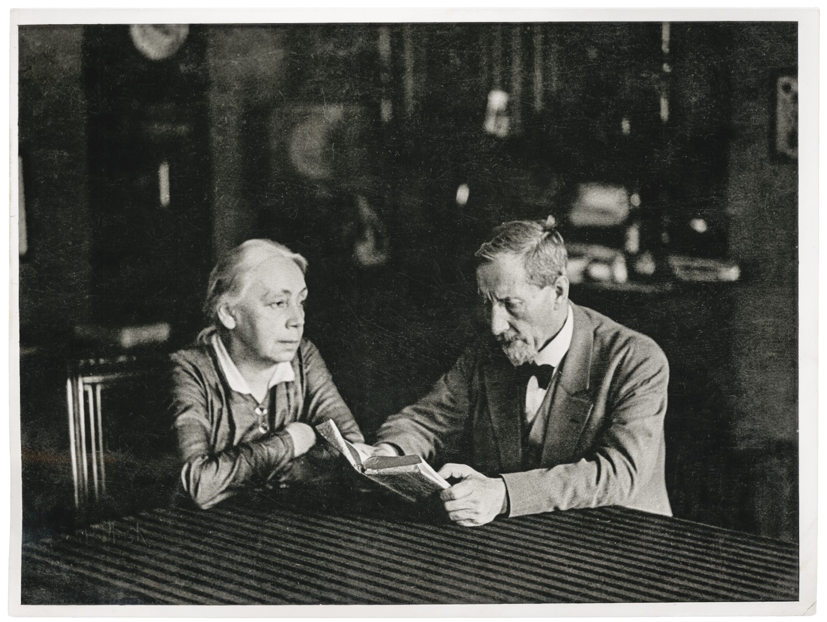 Das Schwarz-Weiß-Foto von Erich Salomon aus dem Jahr 1931 zeigt die Künstlerin Käthe Kollwitz und ihren Ehemann Karl in einem bürgerlichen Wohnzimmer an einem Tisch sitzend. Karl, ein mit Bart und Anzug, hält ein kleines Buch in den Händen und scheint daraus vorzulesen. Käthe sitzt ihm gegenüber, hat ihre Arme auf der Tischplatte verschränkt und blickt ihren Mann dabei aufmerksam und ruhig an.