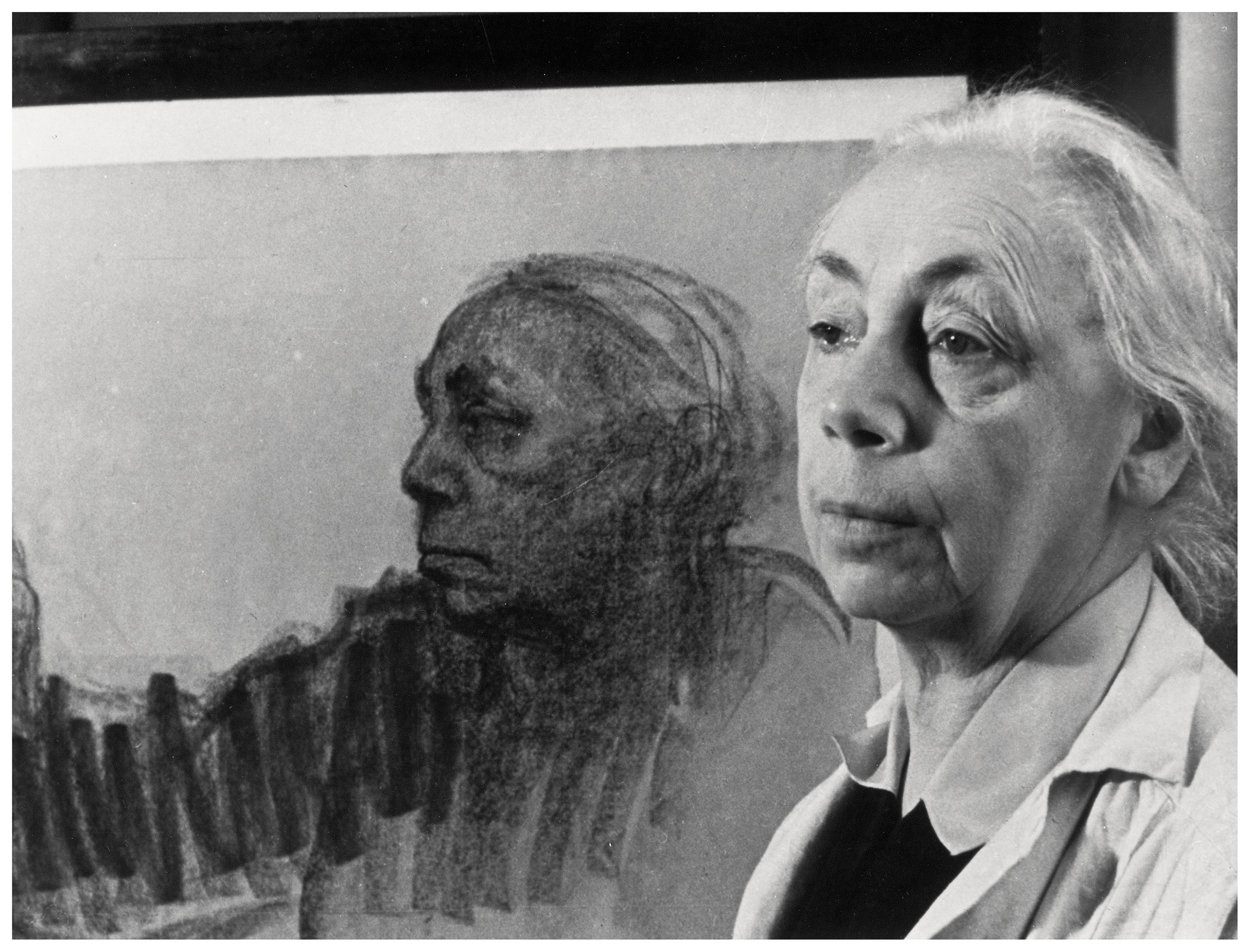 Das querformatige Schwarz-Weiß-Foto zeigt Käthe Kollwitz im rechten Vordergrund, während links hinter ihr eine lebensgroße Zeichnung ihres eigenen Gesichts im Profil zu sehen ist. Sie trägt eine helle Arbeitsjacke über dunkler Kleidung und blickt mit ernstem, nachdenklichem Ausdruck leicht am Betrachter vorbei.