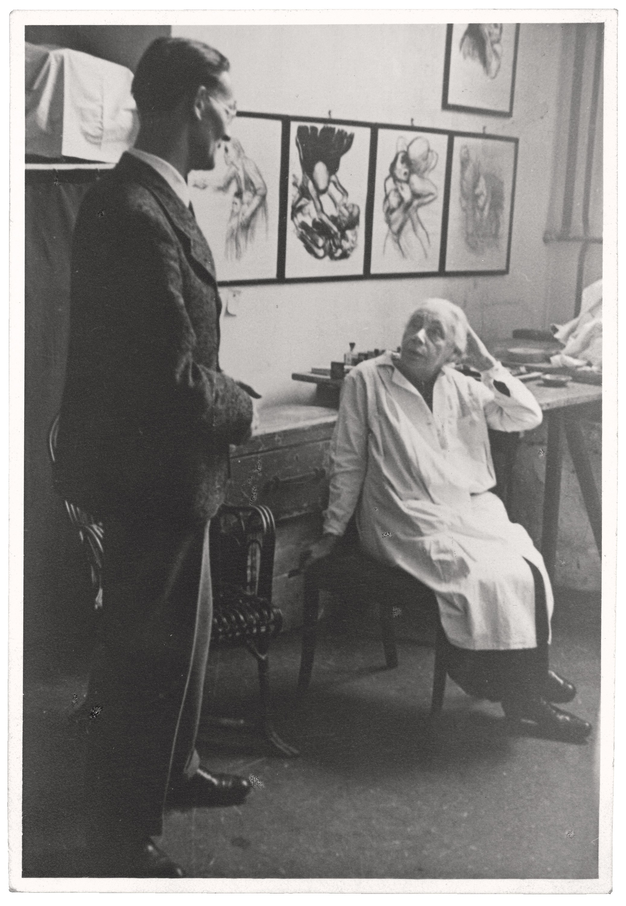 Dieses Schwarz-Weiß-Foto zeigt die Künstlerin Käthe Kollwitz in ihrem Atelier, wie sie im Profil sitzend zu einem vor ihr stehenden Mann aufblickt. Sie trägt einen hellen Arbeitskittel, hat die linke Hand nachdenklich an den Kopf gelehnt und wirkt in ein tiefes Gespräch vertieft. Im Hintergrund hängen an der hellen Wand mehrere ihrer Grafiken aus der Folge »Tod«, dunkel gerahmt.