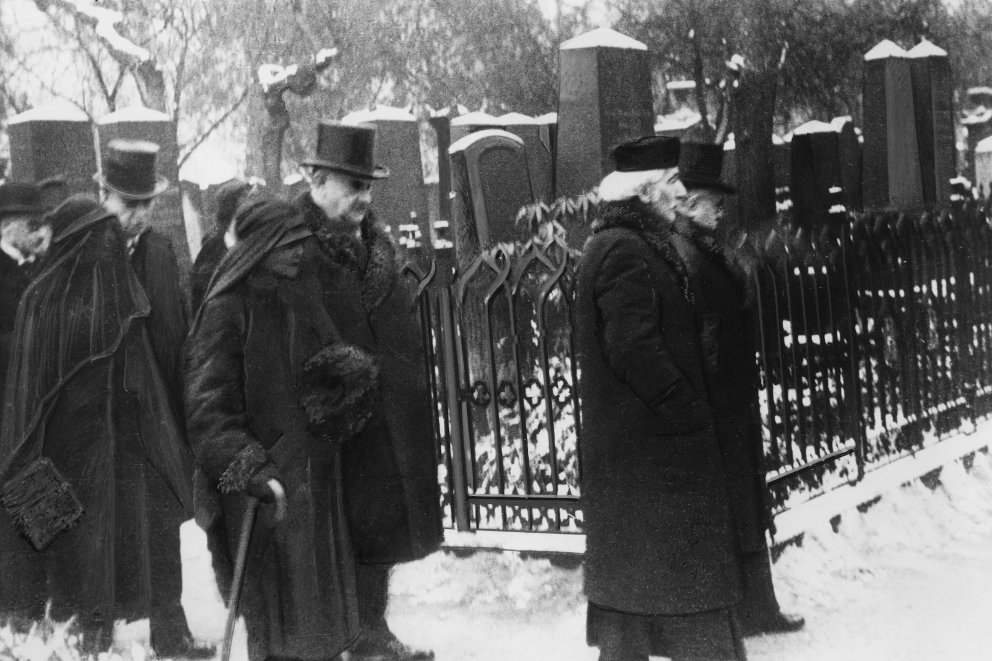 Das historische Schwarz-Weiß-Foto zeigt eine Gruppe von Trauergästen in dunkler Winterkleidung, die bei Schnee an einem kunstvoll verzierten Eisengitterzaun eines Friedhofs entlanggeht. Im Vordergrund sind mehrere Personen in langen Mänteln und Hüten zu sehen, darunter eine Frau mit Gehstock und ein Mann mit Zylinder, während im Hintergrund massive, schneebedeckte Grabsteine aufragen.