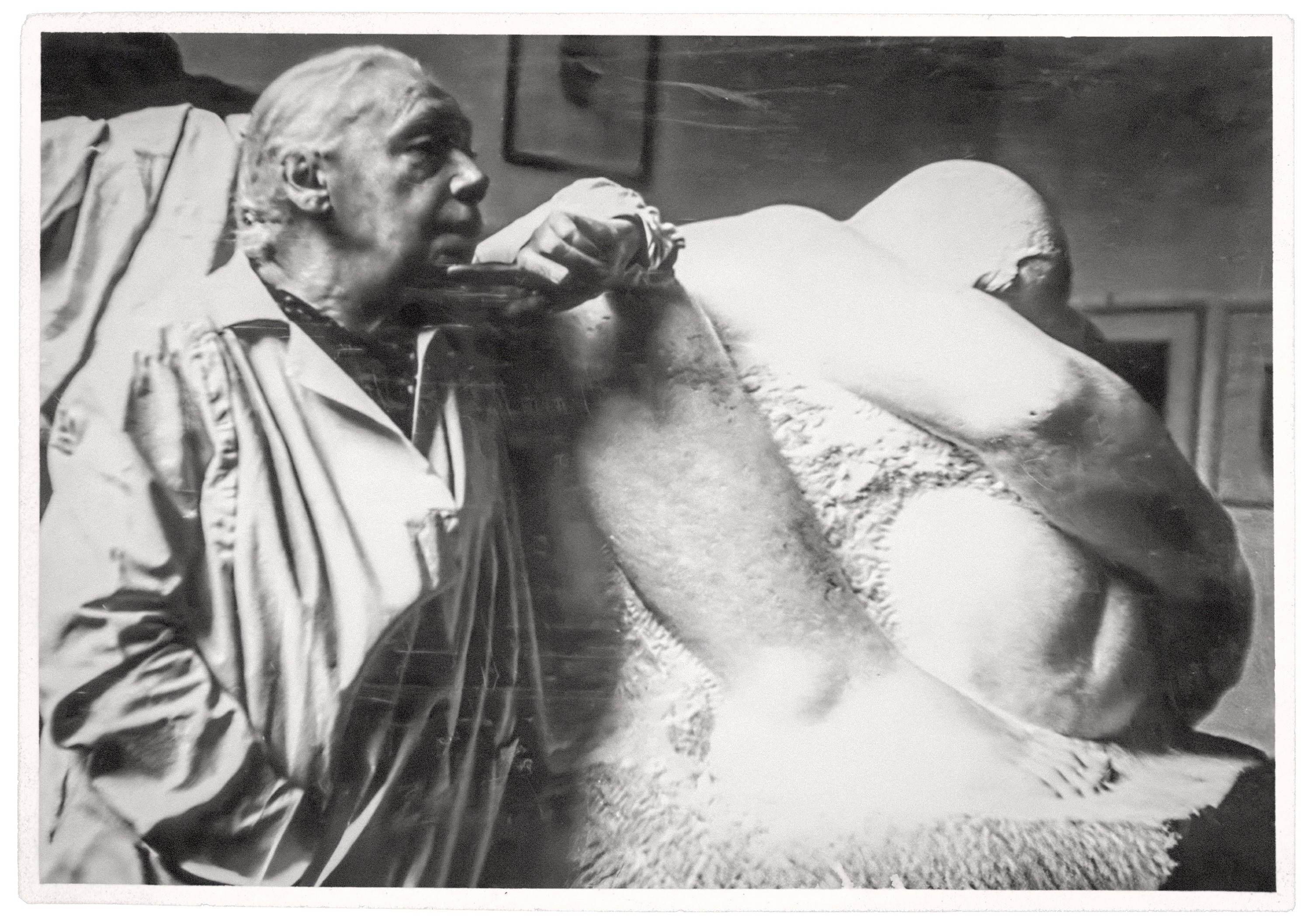 Ein Schwarz-Weiß-Foto zeigt die Bildhauerin Käthe Kollwitz neben ihrer Skulptur »Mutter mit zwei Kindern« in ihrem Atelier. Die Künstlerin steht links im hellen Kittel und stützt ihren Ellebogen auf das Bein der Mutterfigur, während rechts die massiv wirkende, in sich zusammengesunkene Steinfigur den Bildraum dominiert. 