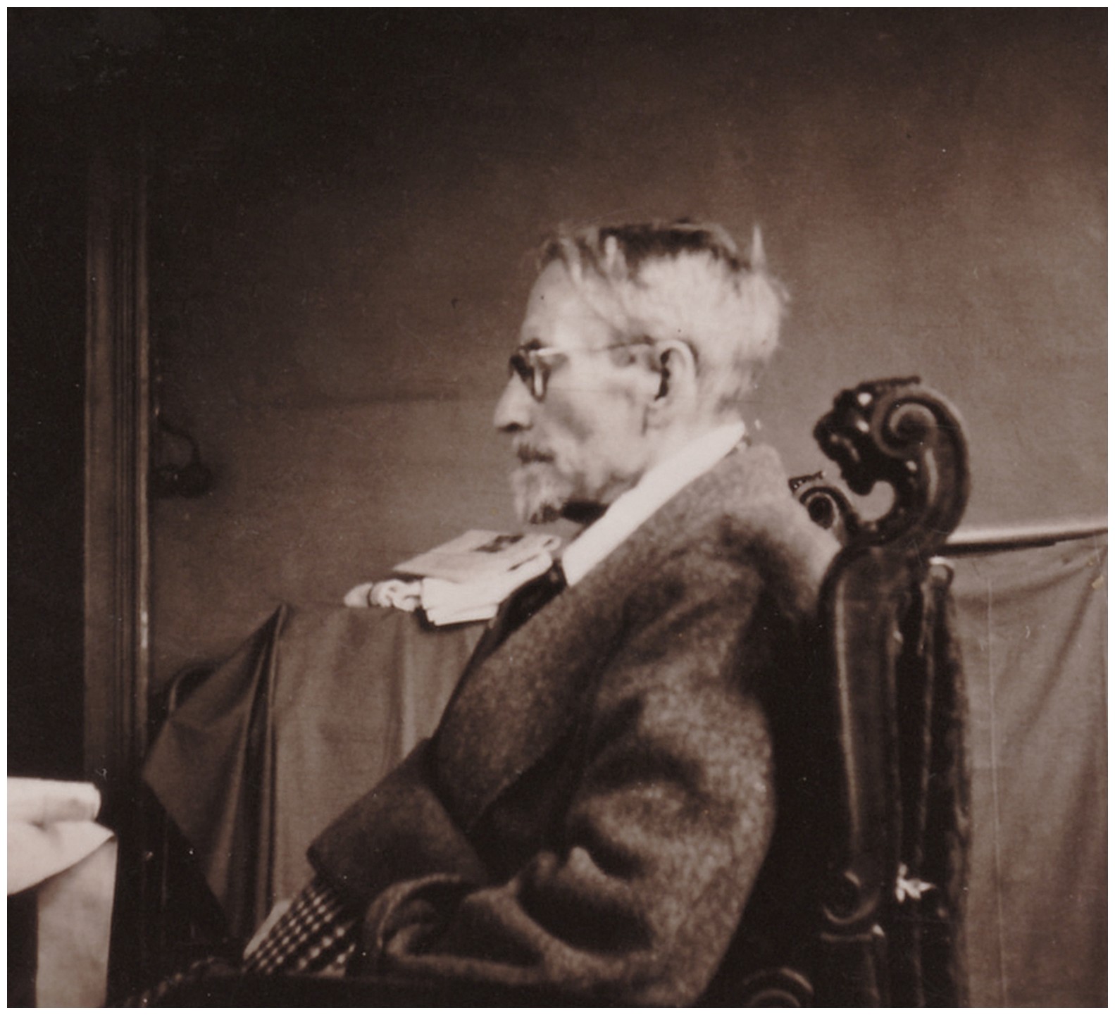Ein Schwarz-Weiß-Foto zeigt das Porträt von Karl Kollwitz als älteren, bärtigen Mann mit Brille im Profil, der in einem dekorativ geschnitzten Holzstuhl sitzt. Er trägt ein dunkles Sakko über einem hellen Hemd und blickt nach links, während im Hintergrund ein Tisch mit Papieren und ein dunkler Vorhang zu erkennen sind.