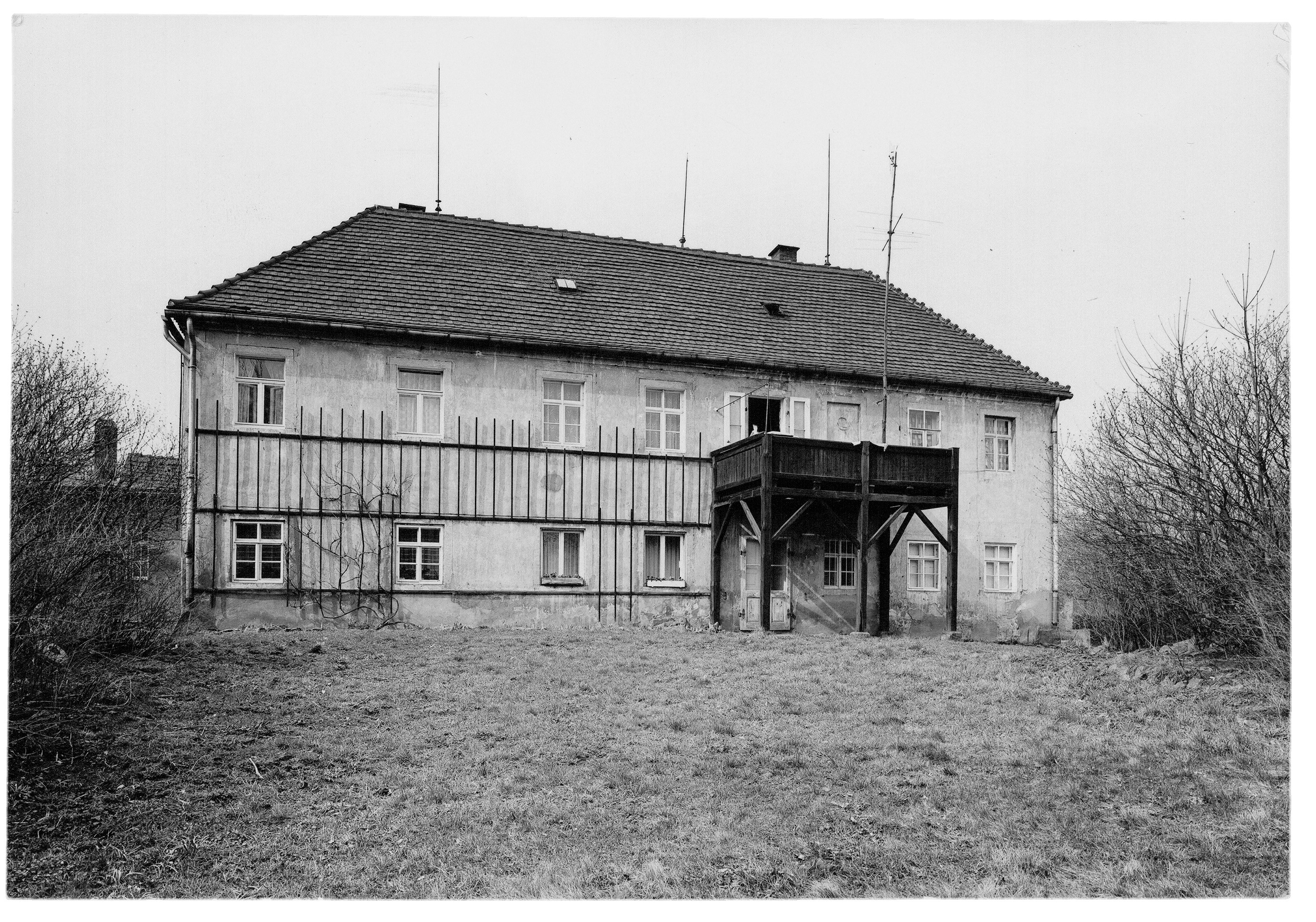 Das Schwarz-Weiß-Foto zeigt die Rückansicht eines großen, zweistöckigen Wohnhauses mit einem hohen Walmdach auf einem weitläufigen Wiesengrundstück. Auffällig ist ein massiver, dunkler Holzbalkon in der Mitte der Fassade, der auf hohen Pfosten über dem Eingangsbereich ruht. Die helle Putzfassade ist durch ein horizontales Holzgerüst und zahlreiche gleichmäßig angeordnete Sprossenfenster streng gegliedert.