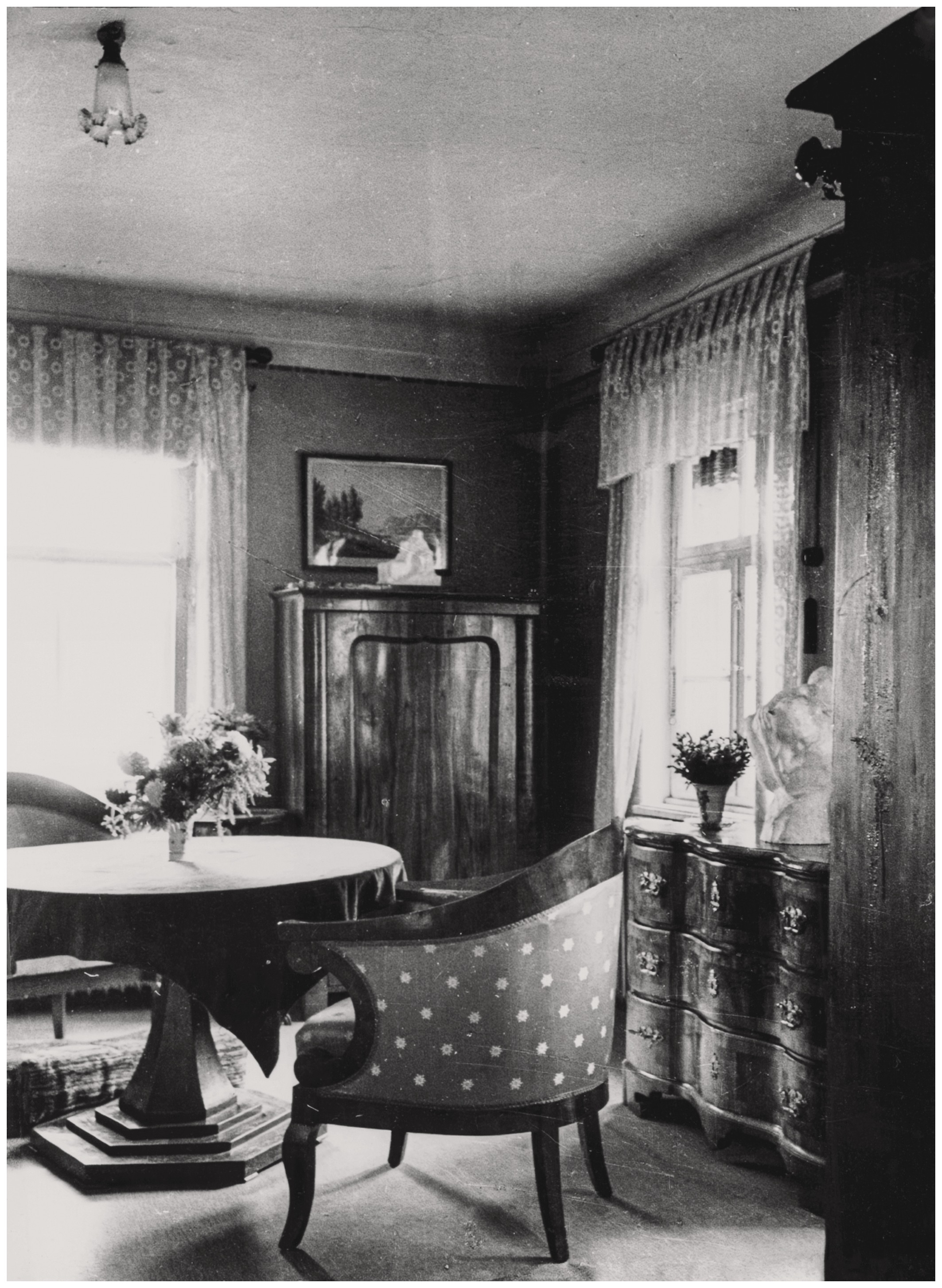 Dieses Schwarz-weiß-Foto zeigt ein behaglich eingerichtetes Zimmer mit einem runden Tisch in der Mitte, der von gepolsterten Sesseln mit einem Punktmuster umgeben ist. Im Hintergrund stehen dunkle, klassische Holzmöbel wie eine geschwungene Kommode und ein Schrank, während helle Spitzengardinen an den Fenstern den Raum mit weichem Licht füllen. Auf den Möbeln sind Vasen mit Blumen und kleine Skulpturen verteilt, was dem Raum eine persönliche und museale Atmosphäre verleiht: Es sind die von Kollwitz angefertigen Gipsmodellen von »Zwei wartende Soldatenfrauen« auf dem Schrank und »Mutter mit Kind über der Schulter« auf der Kommode rechts. 