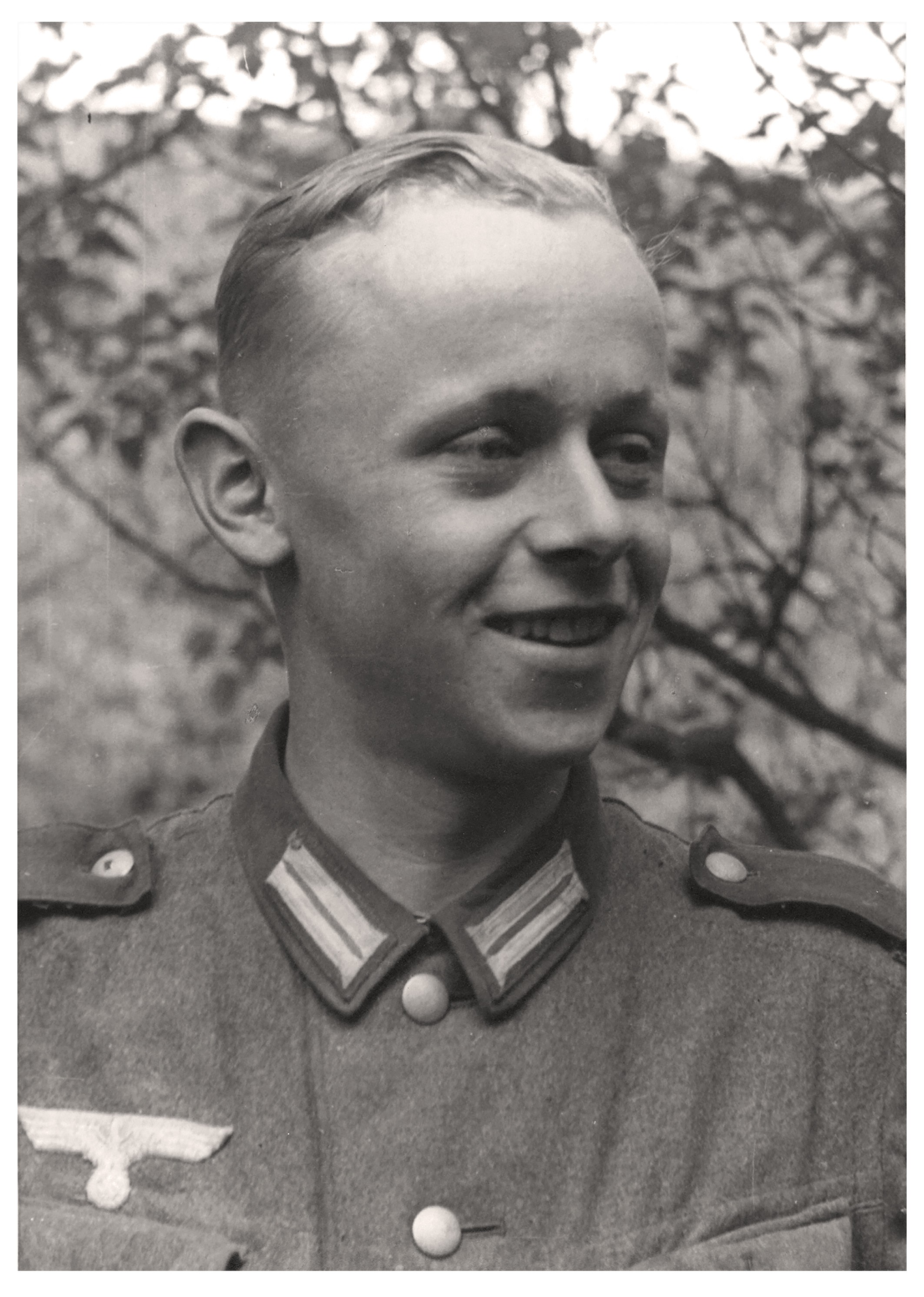 Das Schwarz-Weiß-Porträt aus dem Jahr 1941 zeigt einen jungen, lächelnden Mann in einer deutschen Wehrmachtsuniform aus der Zeit des Zweiten Weltkriegs. Er trägt eine Feldbluse mit markanten Kragenspiegeln und dem Hoheitsabzeichen über der rechten Brusttasche, während sein Blick zur Seite gerichtet ist. Sein helles Haar ist ordentlich zur Seite gekämmt und der Hintergrund besteht aus leicht unscharfen Blättern und Zweigen im Freien.