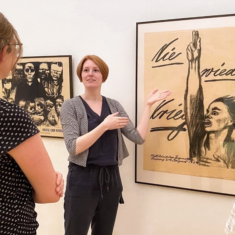 In hellen Ausstellungsräumen des Kollwitz Museum Köln erklärt eine Kunstvermittlerin mit ausladenden Handbewegungen zwei aufmerksam zuhörenden Besucherinnen das berühmte Antikriegsplakat "Nie wieder Krieg" von Käthe Kollwitz. Das Werk steht im Fokus und zeigt die ausdrucksstarke, schwarz-weiße Kohlezeichnung einer Person, die mit weit aufgerissenem Mund und einer zum Schwur erhobenen Hand leidenschaftlich „Nie wieder Krieg“ fordert.