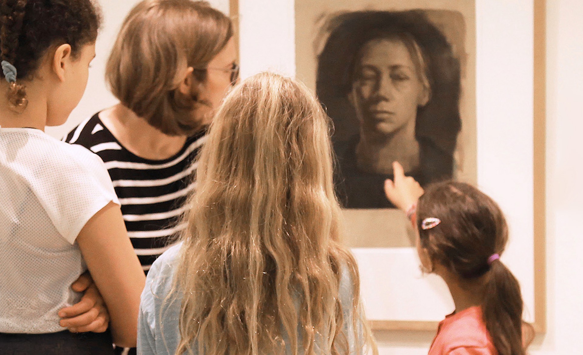 Das Bild zeigt eine Frau und drei junge Mädchen, die in einem Museum dicht vor einem großen, gerahmten Porträt stehen. Während die Kunstvermittlerin und zwei der Kinder das Werk aufmerksam betrachten, deutet ein Mädchen mit dem Finger direkt auf das Gesicht des Selbstbildnisses von Käthe Kollwitz.