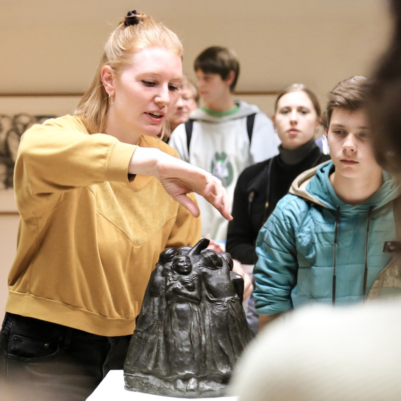 Ein Foto zeigt eine Kunstvermittlerin im Kollwitz Museum Köln. Sie trägt ein senfgelbes Sweatshirt neigt sich erklärend über die kleine, dunkle Bronzeplastik, den  „Turm der Mütter“ von Käthe Kollwitz, die auf einem weißen Sockel steht. Sie ist umringt von einer Schülergruppe , die aufmerksam die kreisförmig angeordneten, schützenden Frauenfiguren der Skulptur betrachten und den Ausführungen zuhören.