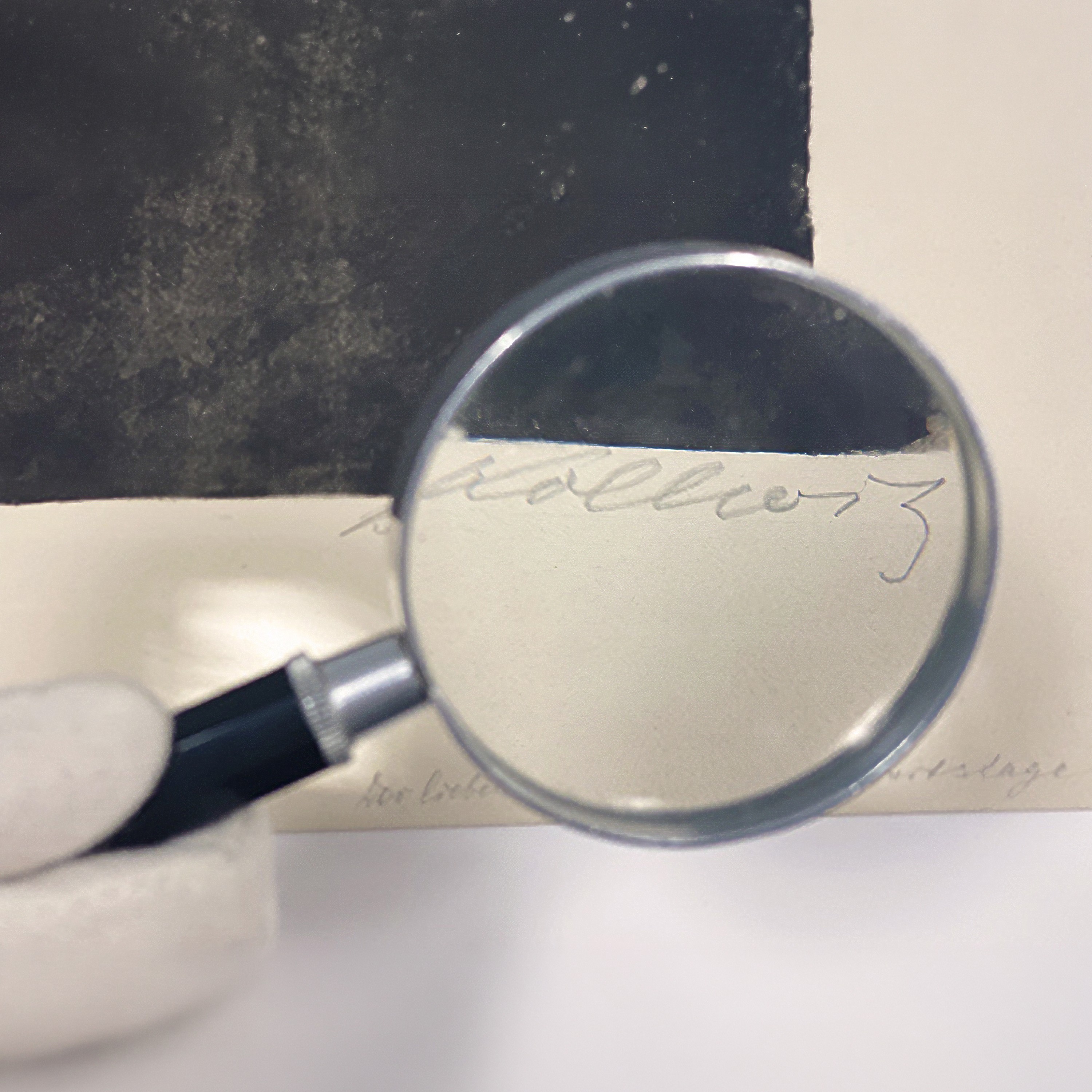 The picture shows a magnifying glass being held by a hand wearing a white glove over the handwritten signature ‘Kollwitz’ on a light-coloured margin of paper. Thanks to the magnification, the artist’s signature is clearly legible beneath a dark, textured artwork.