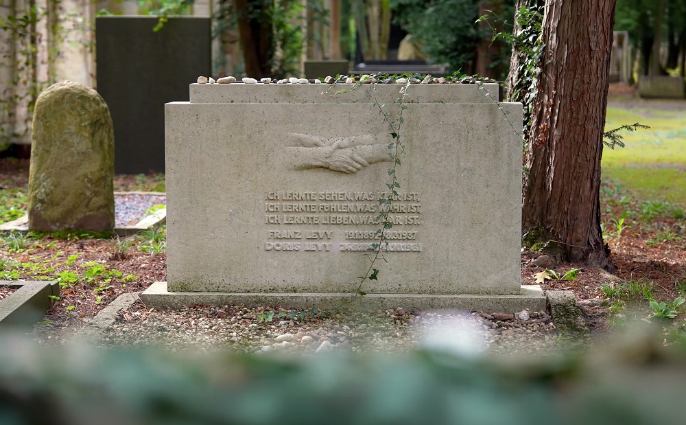 The photograph shows the tomb of the Levy family in the Jewish cemetery of Cologne-Bocklemünd: an upright white marble stone measuring 80 × 150 cm, bearing the relief of four clasping hands by Käthe Kollwitz. Beneath the relief is the inscription: »Ich lernte sehen, was klar ist / ich lernte fühlen, was wahr ist / ich lernte lieben, was rar ist« (I learned to see what is clear / I learned to feel what is true / I learned to love what is rare).