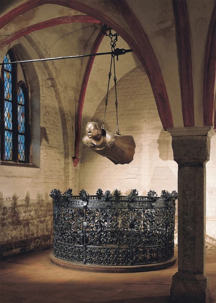 The Picture by the photographer Uwe Seemann shows the bronze sculpture »Der Schwebende« (Floating Angel) by Ernst Barlach — the third cast of 1952 after the Cologne example — in its present location, the originally intended place in the eastern bay of the north aisle in Güstrow Cathedral. The life-size bronze figure, hovering horizontally, hangs freely in the air from two chains, its straight body and arms crossed over the chest radiating a deep serenity. The face of the sculpture has closed eyes and seems absorbed in itself, while the flowing form of the robe reinforces the impression of a gentle flight. Beneath the »Schwebender« stands a circular, ornately decorated wrought-iron grille, which further anchors the figure in the space and marks a place of remembrance.