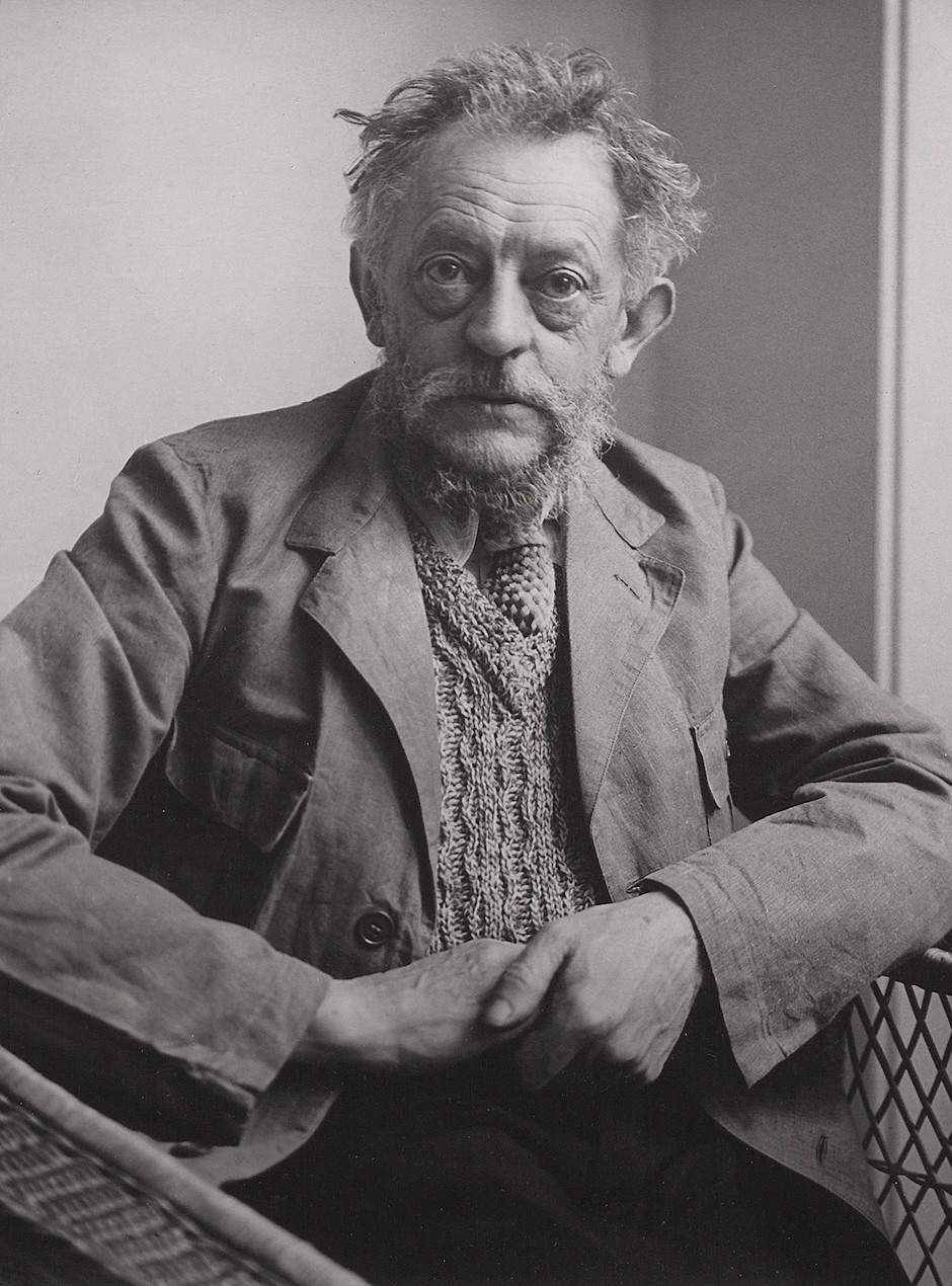 The black-and-white photograph shows a head-and-shoulders portrait of the sculptor Ernst Barlach in a frontal view, taken in 1933 by Hildegard Heise. He is an elderly man with tousled, light-coloured hair, a thick, grey-streaked full beard and an intense gaze directed straight at the camera. Barlach is wearing a coarse, light-coloured jacket over a distinctive knitted waistcoat with a tie, and is holding his hands above his lap, his elbows resting on the armrests of his wicker armchair.