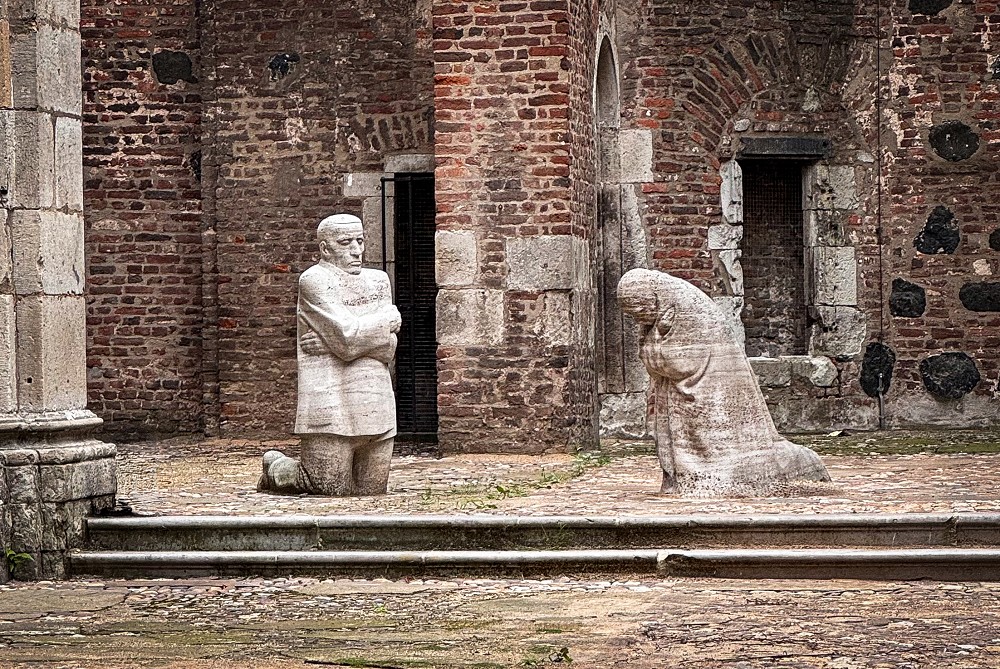 This photograph by Christian Nitz shows Käthe Kollwitz’s Memorial »Grieving Parents«, which is situated in the open interior of the ruins of the Old St Alban’s Church in Cologne. Amidst the weathered brick walls, two life-size, block-like stone figures kneel facing each other in silent grief: on the left, the father stands motionless, his gaze fixed straight ahead and his arms crossed tightly across his chest, whilst on the right, the mother, deeply bowed and lost in her own thoughts, mourns their son.