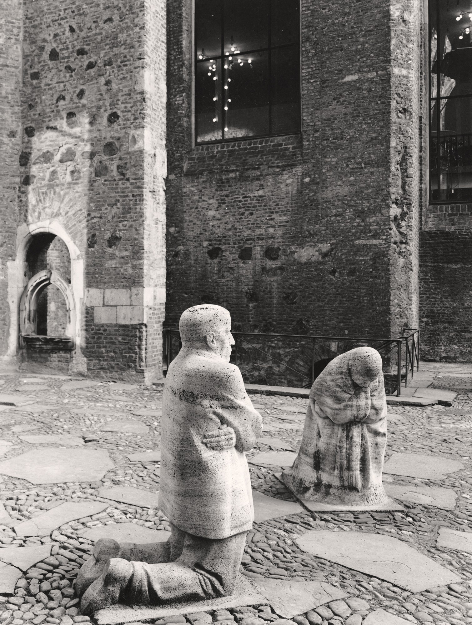 Lothar Schnepf’s black-and-white photograph from 1999 shows replicas of Käthe Kollwitz’s memorial ‘Grieving Parents’ in the ruins of the Old St Alban’s Church in Cologne, the first federal memorial to those who died in the two world wars. The figures – the father, seen in profile, and the mother, facing the viewer – are set into the paved floor. In the background are the brick walls of the church with a large window offering a view into the neighbouring Gürzenich banquet hall.