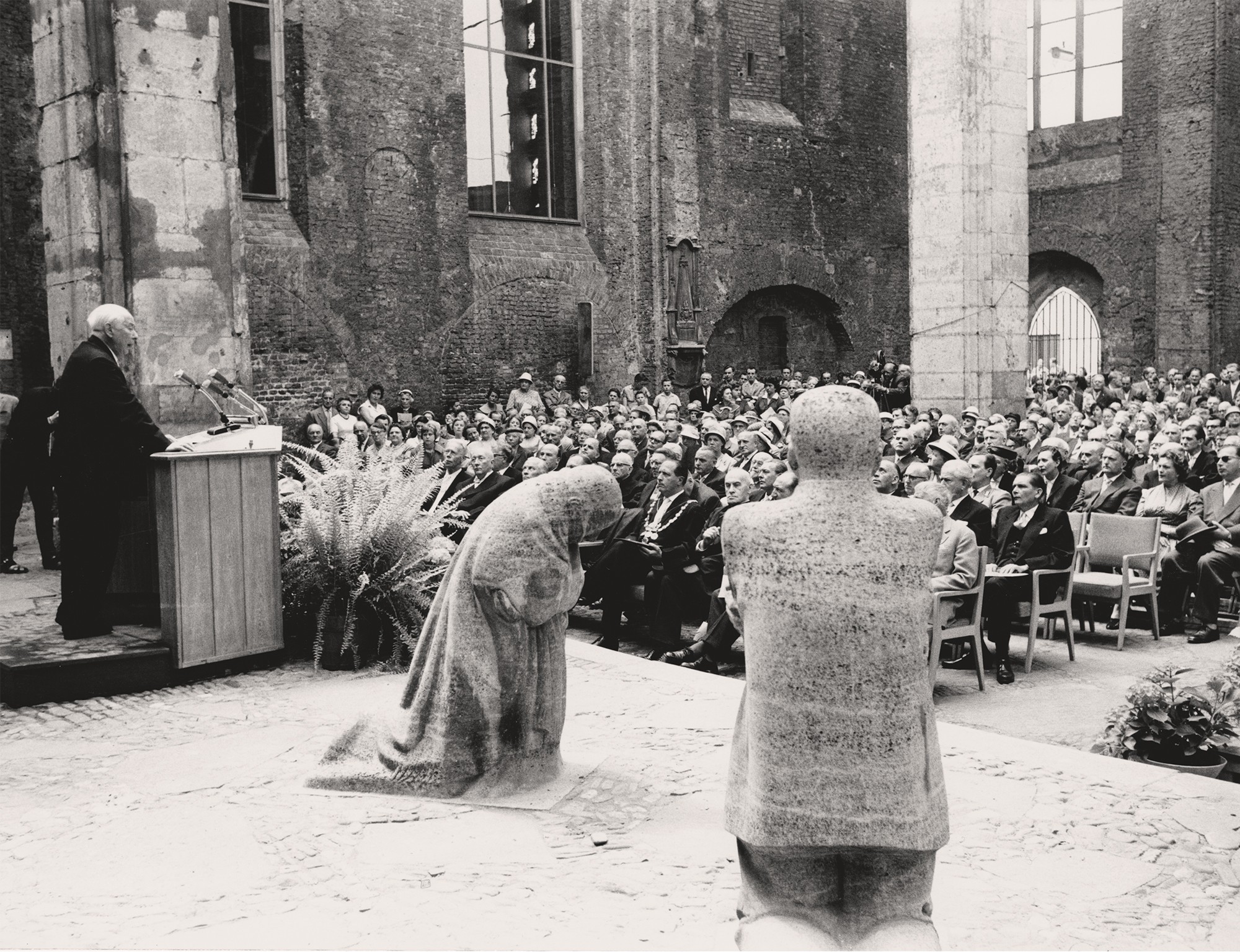 A black-and-white photograph by Hansherbert Wirtz shows a view from the choir of the ruins of the Old St Alban’s Church in Cologne, looking out towards the congregation during its inauguration as a federal memorial. The image shows the two stone sculptures ‘Grieving Parents’ – the father on the left, the mother in the centre – with Federal President Theodor Heuss on the right-hand edge of the frame as he delivers his speech.
