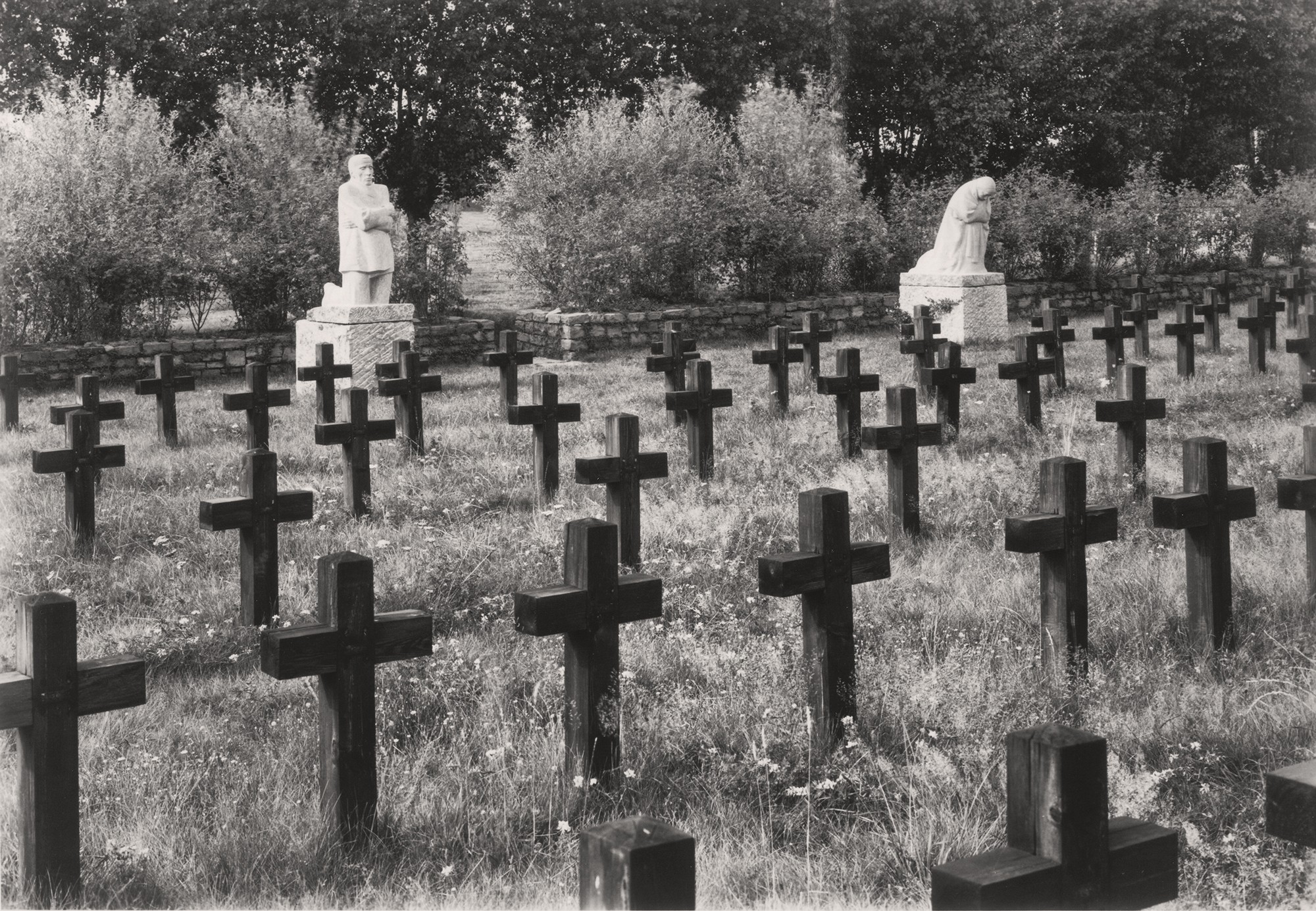 This black-and-white photograph by Willi Moegle, taken in 1937, shows the pair of sculptures »Grieving Parents« in their original setting at the Roggeveld Military Cemetery in Belgium, where they stood from July 1932 until 1955. Viewed from the right, a summer meadow dotted with numerous wooden crosses can be seen in the foreground; in the background, set against a low rubble-stone wall, stand the sculptures – the father on the right, the mother on the left – with the stone wall opening up between them to form a narrow exit from the cemetery.