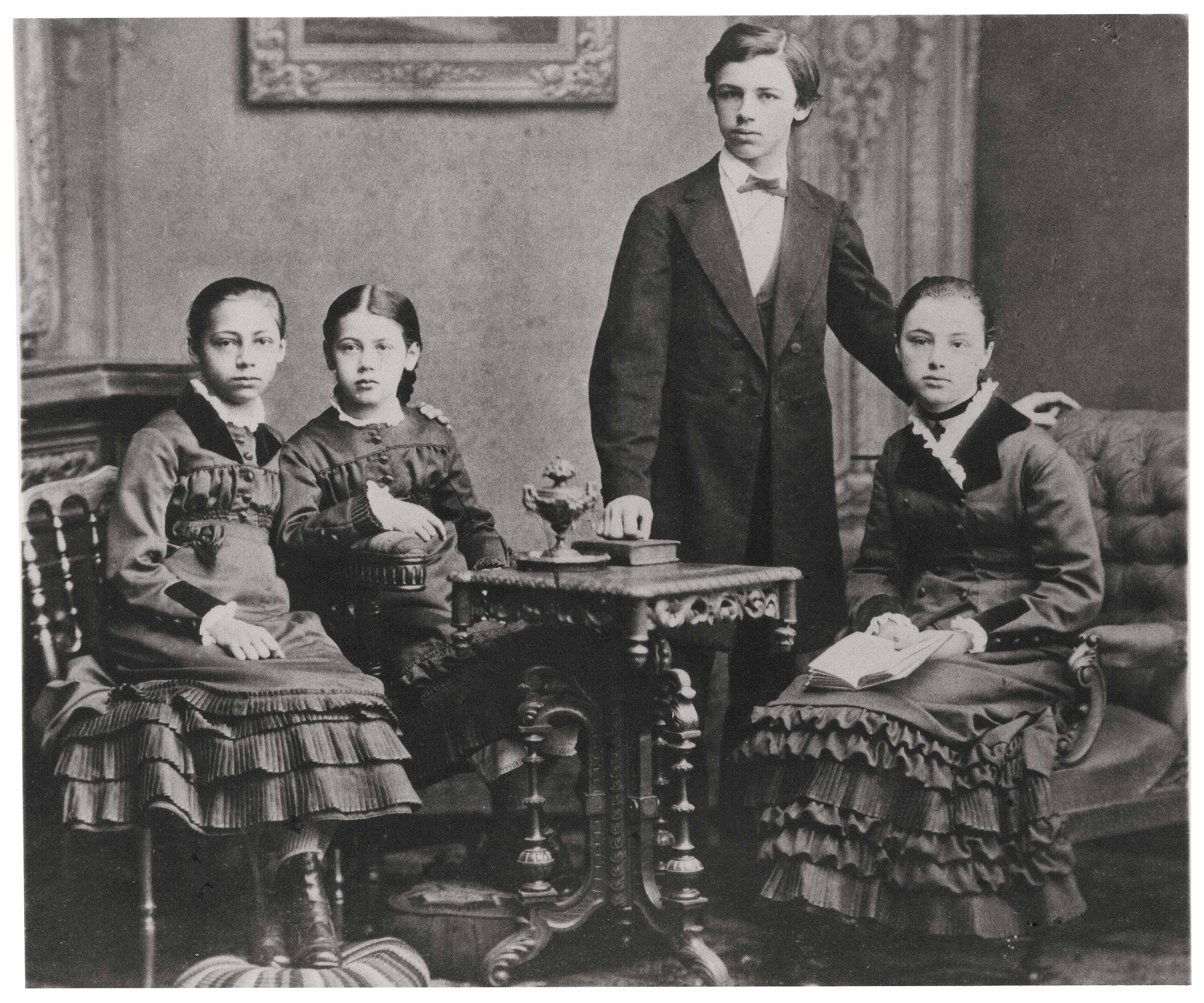 The image is a black-and-white photograph dating from around 1880, showing the four well-dressed Schmidt siblings—Käthe, Lisbeth, Konrad, and Julie, from left to right—in a middle-class parlour. At the centre stands Konrad, dressed in a dark suit with a white shirt and bow tie, surrounded by the three girls in dark dresses with prominent ruffles and white collars. The group poses solemnly and motionlessly around an ornately carved wooden table.