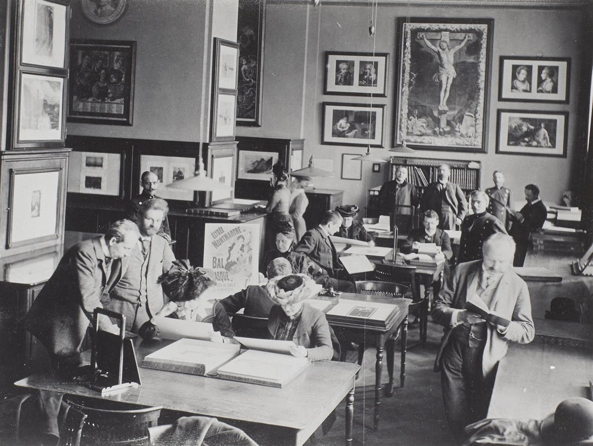 The photograph from 1913 shows the study hall of the Kupferstich-Kabinett (Department of Prints and Drawings) in Gottfried Semper's museum building — a busy, historical reading room or study collection in which numerous people in formal dress work intently over portfolios and books at large wooden tables. The walls are hung densely with framed prints, drawings and a large crucifix painting, underscoring the atmosphere of a scholarly institution or art collection. In the foreground, men and women stand and sit in period dress, such as jackets and broad-brimmed hats, while the incoming light creates a contrast-rich, calm working mood. In the background to the right is Max Lehrs.