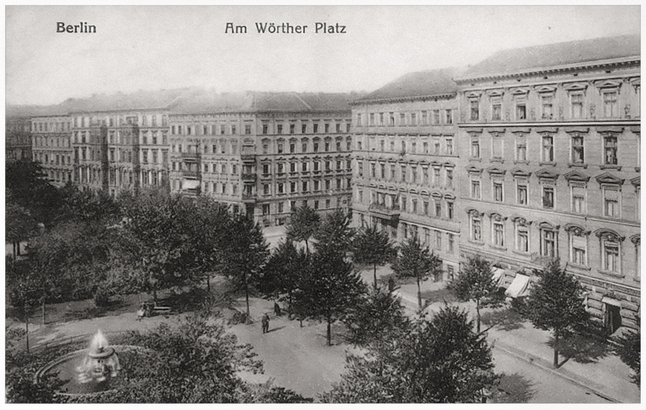 The image shows a historic black-and-white postcard of Wörther Platz in Berlin, depicting a lively urban square with a central fountain and dense tree cover. In the background rise impressive multi-storey residential buildings with richly ornamented façades, characteristic of Berlin’s Gründerzeit architecture. The scene conveys a peaceful atmosphere and captures the character of urban life in a bygone era.