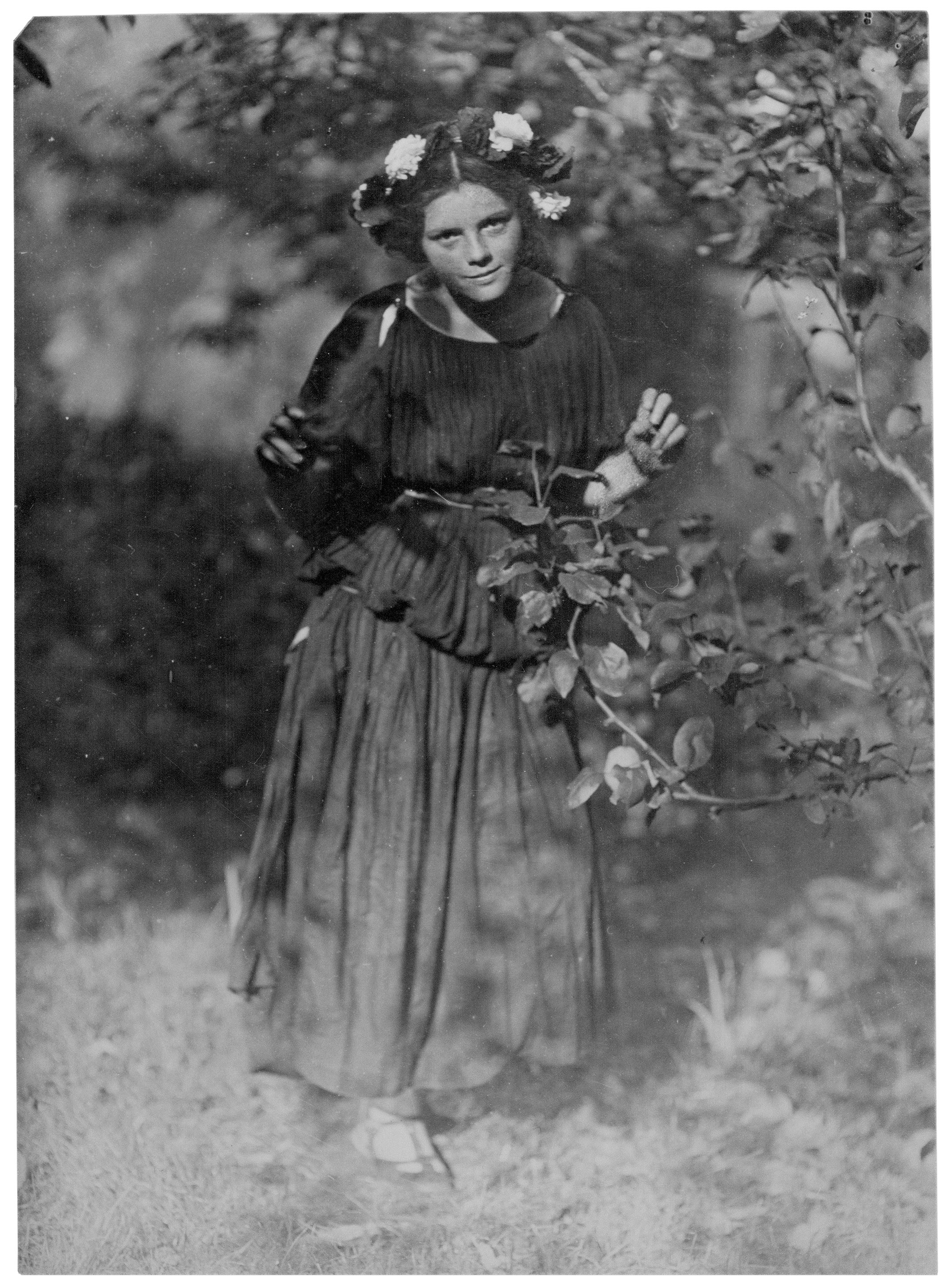 The historic black-and-white photograph, dating from 1912, shows Hanna Stern (1896–1988), later known as Johanna Hofer, as a young girl in a natural setting, presumably a garden. She wears a dark, loosely draped garment with a full skirt and a striking floral wreath of light and dark blossoms in her hair. Standing at the centre of the image, she gazes attentively ahead, surrounded by foliage and branches that partially extend into the foreground, softly out of focus.