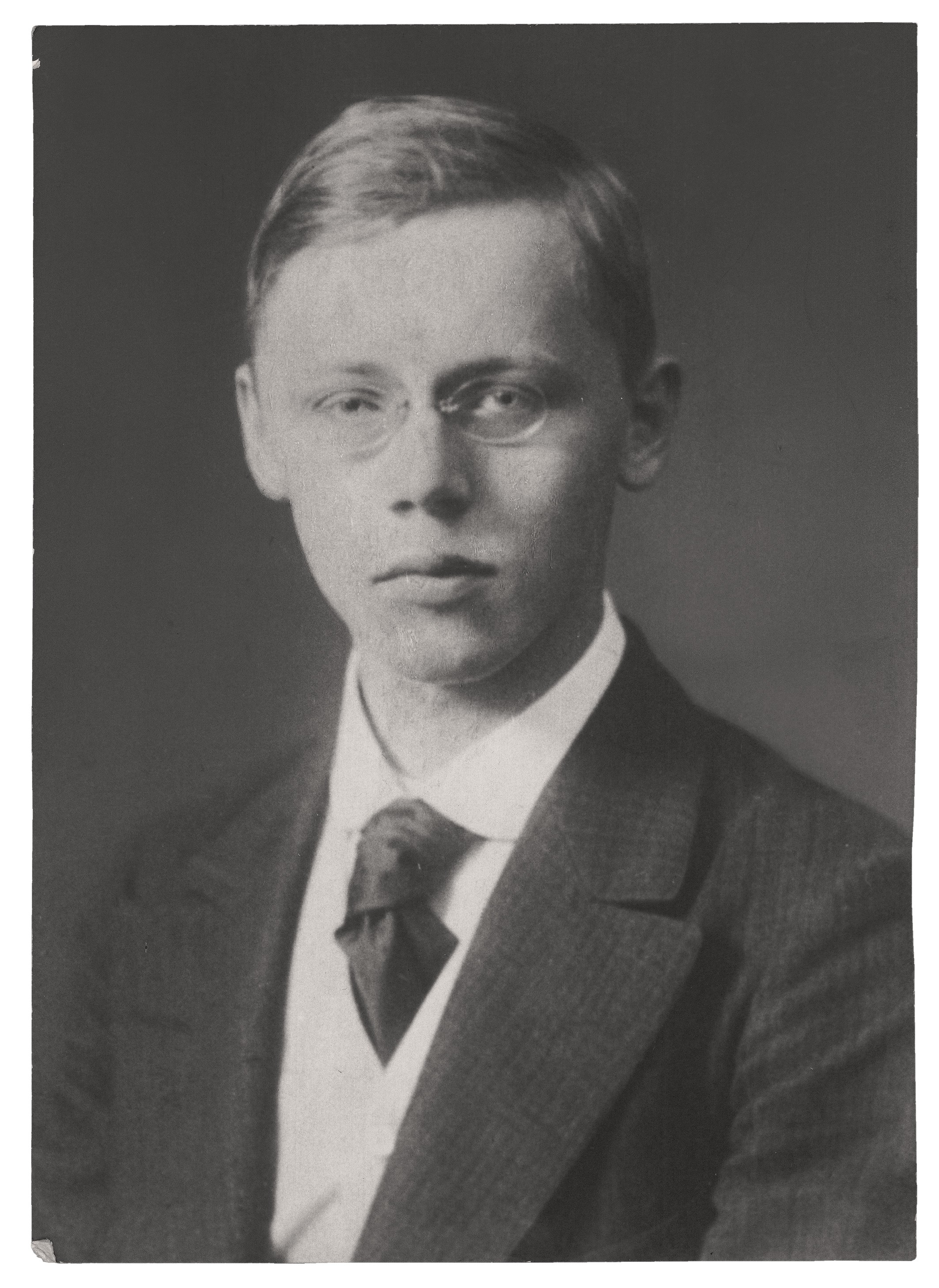 The black-and-white portrait shows a bust-length view of Hans Kollwitz as a student, with a slender face and a serious, somewhat melancholic expression. He wears round spectacles, and his short, light-coloured hair is neatly parted to one side. His attire consists of a dark, finely striped jacket worn over a light waistcoat and a white shirt, complemented by an elaborately knotted dark tie.