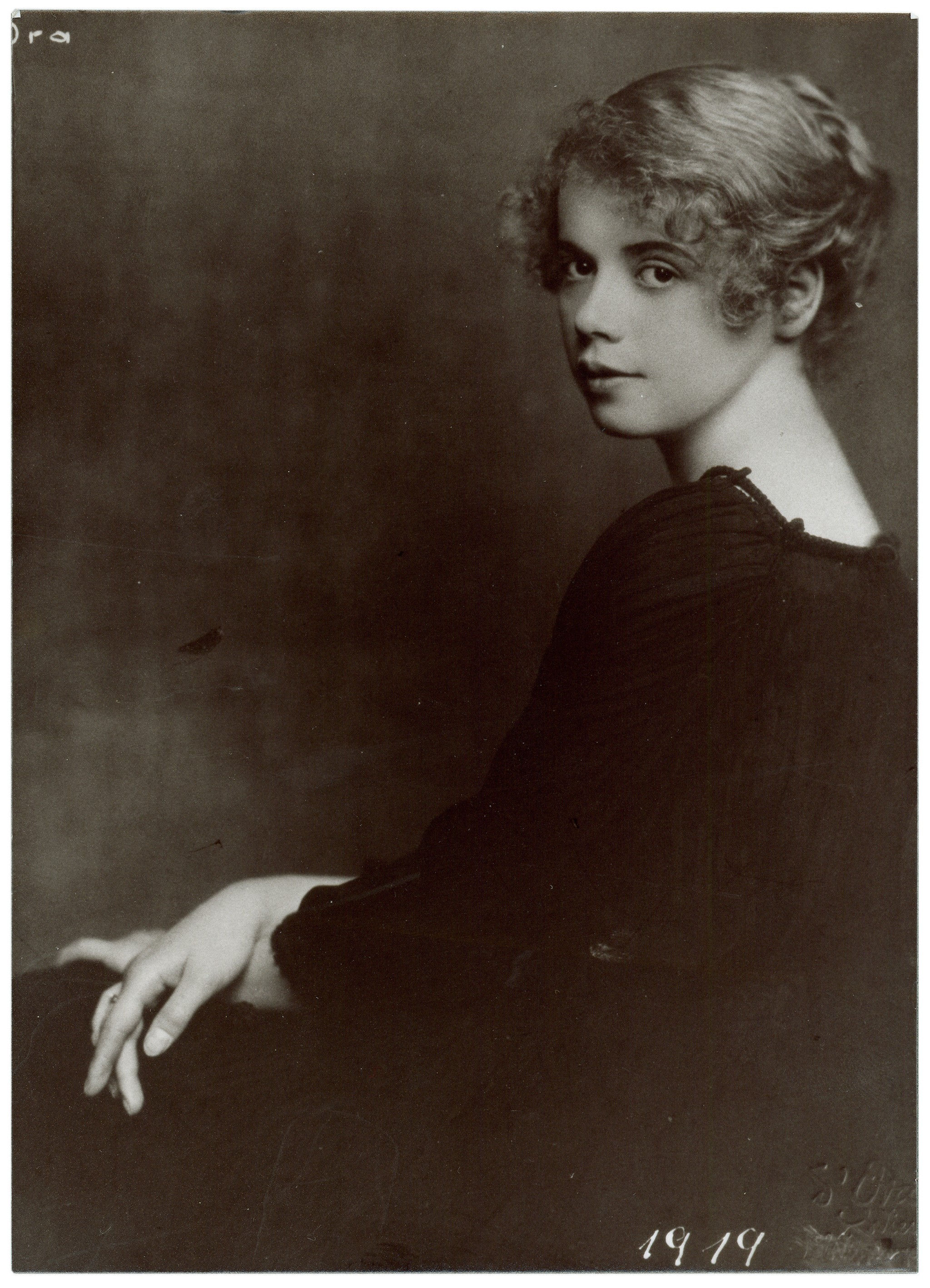 This portrait, dating from 1919, depicts Katharina Stern (1897–1984), also known as Katta Sterna, as a young woman in a classic profile view, gently turning her head over her shoulder towards the viewer. Her light, curly hair is elegantly pinned up, accentuating her delicate facial features and her calm, steady gaze. She wears a dark blouse with subtle gathering at the neckline, which stands out clearly against the softly shaded background.