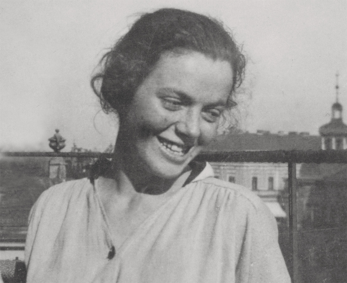 The image shows a black-and-white photograph of Regula Stern (1894–1978) as a young woman, standing before a railing outdoors in a light, loosely flowing top. She looks down with a smile, her head slightly inclined, while her dark, faintly wavy hair is tousled a little by the wind. In the blurred background, the outlines of city buildings and a tower can be made out beneath a bright sky.