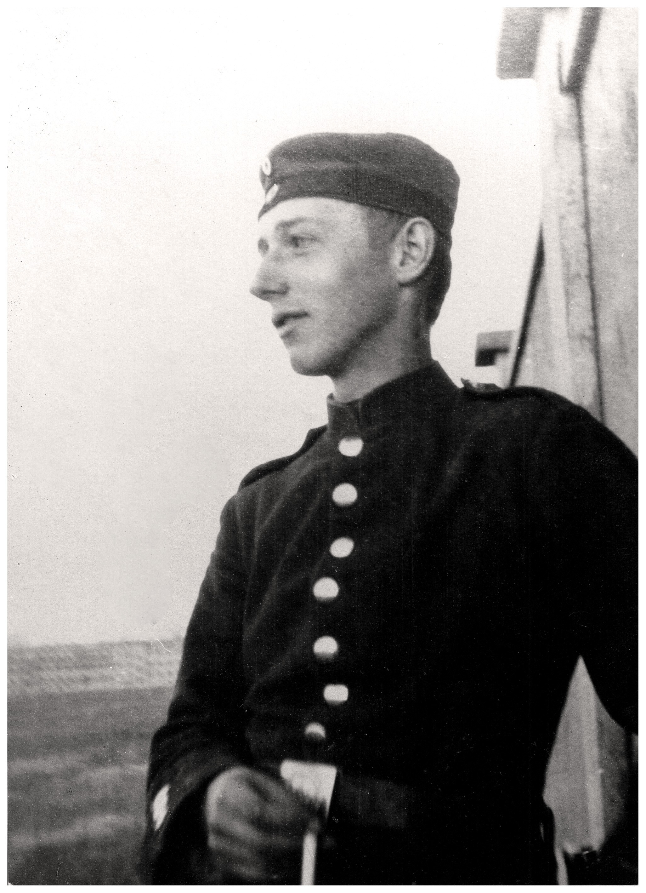 This black-and-white photograph shows Peter Kollwitz as a young man in military uniform, seen in profile. He is looking to the left and wears a dark tunic with a vertical row of light-coloured buttons and a flat field cap with a small round badge at the front. He leans lightly against a wall with one arm, while the background reveals the sky and an open landscape.