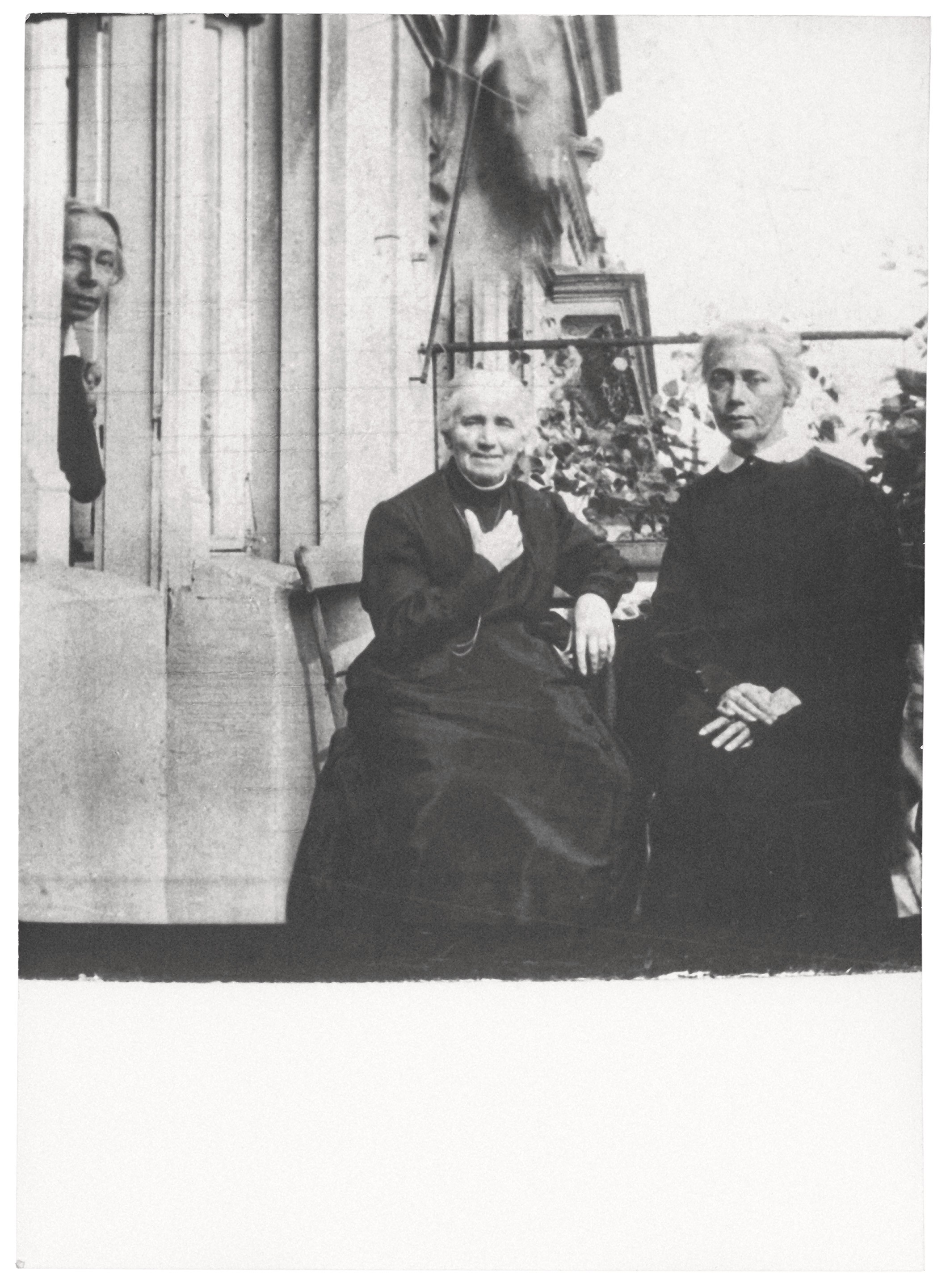 This black-and-white photograph shows three women on a balcony. At the center sits an elderly woman, Katharina Schmidt, with gray hair, dressed in a dark gown, gently resting her hand on her chest. To her right stands her daughter Lise, wearing a white collar and looking on with a stern expression, while to the left in the background, her daughter Käthe peers out from a window. The scene feels calm and intimate, framed by stone columns and a few plants in the background.