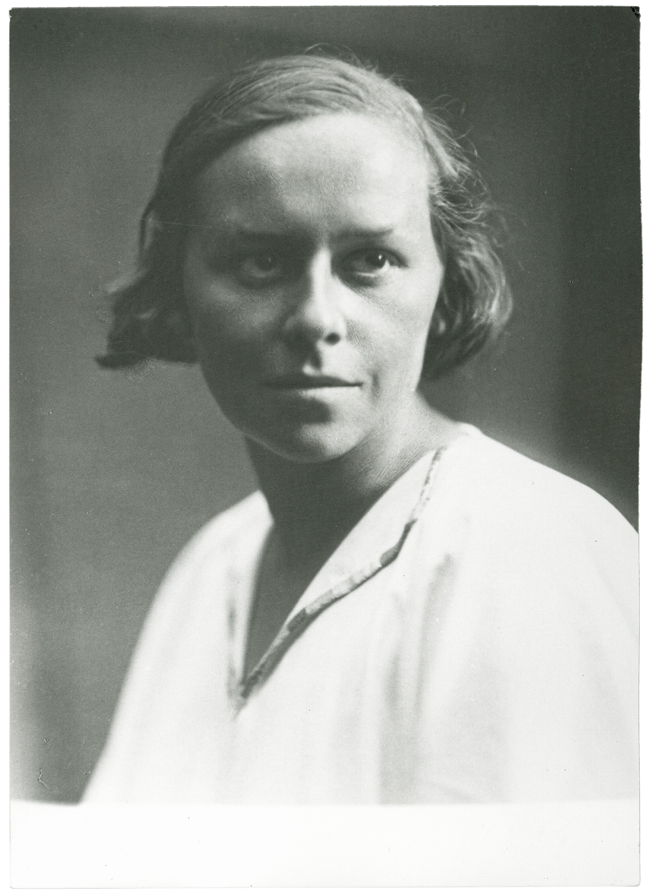 The black-and-white portrait shows a young woman with short, dark hair that is slightly wavy at the sides and almost covers her ears. She is wearing a simple white blouse with a patterned collar and looks directly into the camera with a thoughtful, slightly serious expression. The background is neutral and softly blurred, drawing full attention to her distinct facial features and the gentle side lighting that illuminates her face.