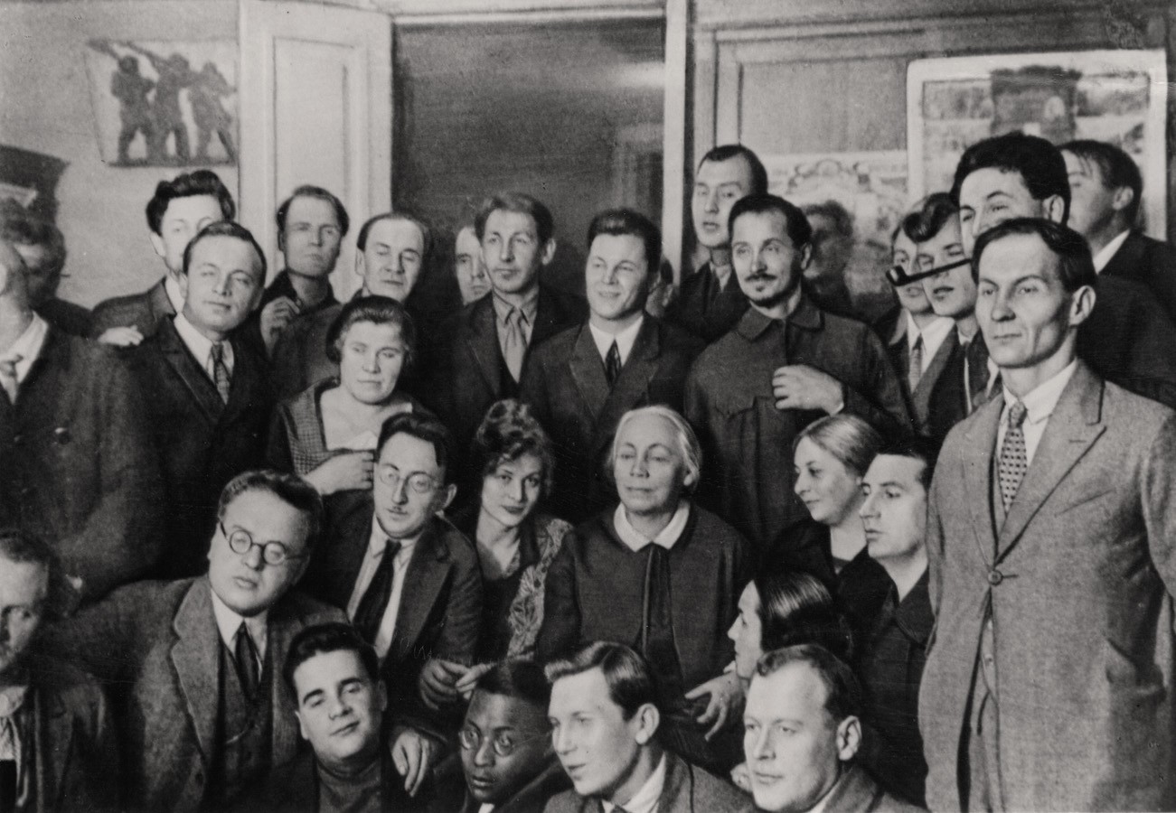 This black-and-white photograph from 1927 shows a large group of around twenty-five people, predominantly men in formal suits, posing closely together for a portrait. At the center of the group sits Käthe Kollwitz, with light-colored hair, dressed in a dark dress with a white collar, while sketches and posters can be seen hanging on the wall in the background. The serious expressions and formal attire give the photograph a dignified and distinctly historical atmosphere.