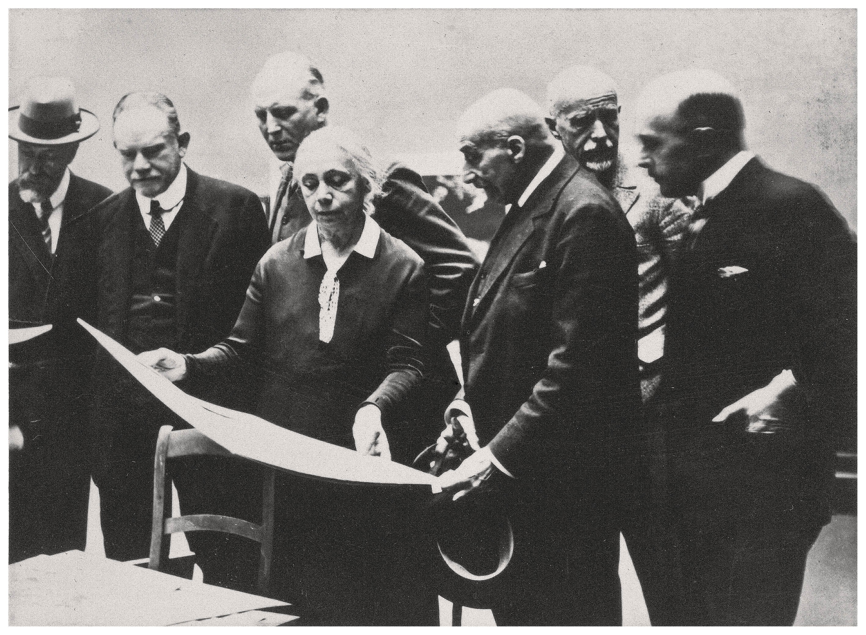 The black-and-white photograph shows a group of six people—five men and one woman—gathered closely around a table, intently examining a large sheet of paper. At the center stands the artist Käthe Kollwitz, holding the sheet, while the men, dressed in dark suits and some wearing hats, stand around her and focus their attention on the document. The scene has a historical and formal character; the serious expressions and close grouping of the figures convey an atmosphere of collaborative work and critical evaluation.