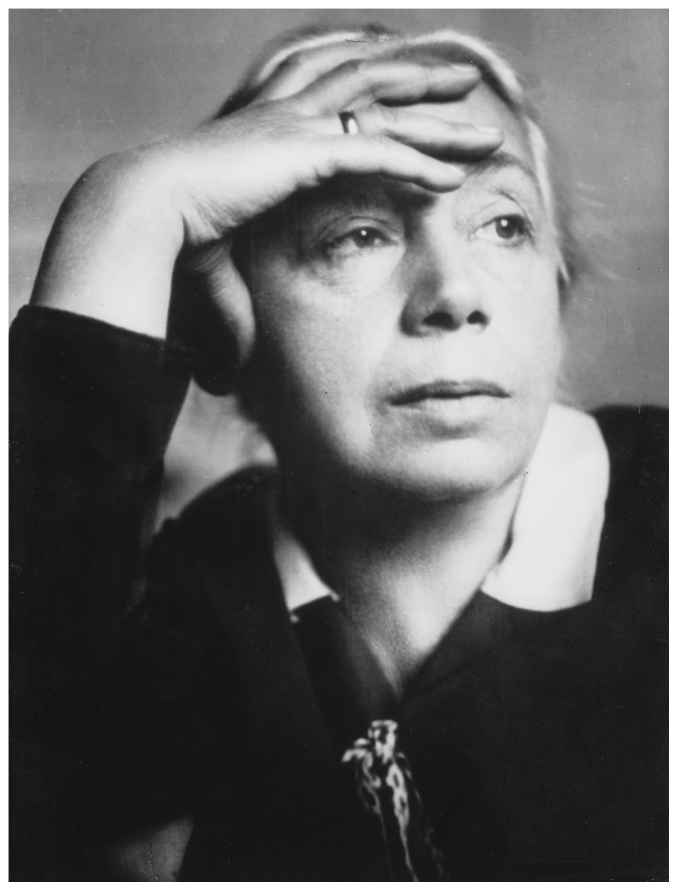 This black-and-white portrait by Emil O. Hoppé from 1928 shows the artist Käthe Kollwitz in a thoughtful and serious pose. She gazes past the viewer into the distance, while her right hand rests flat against her forehead, with a simple ring visible on her finger. Her expression appears focused and slightly melancholic, further emphasized by the interplay of light and shadow across her features.