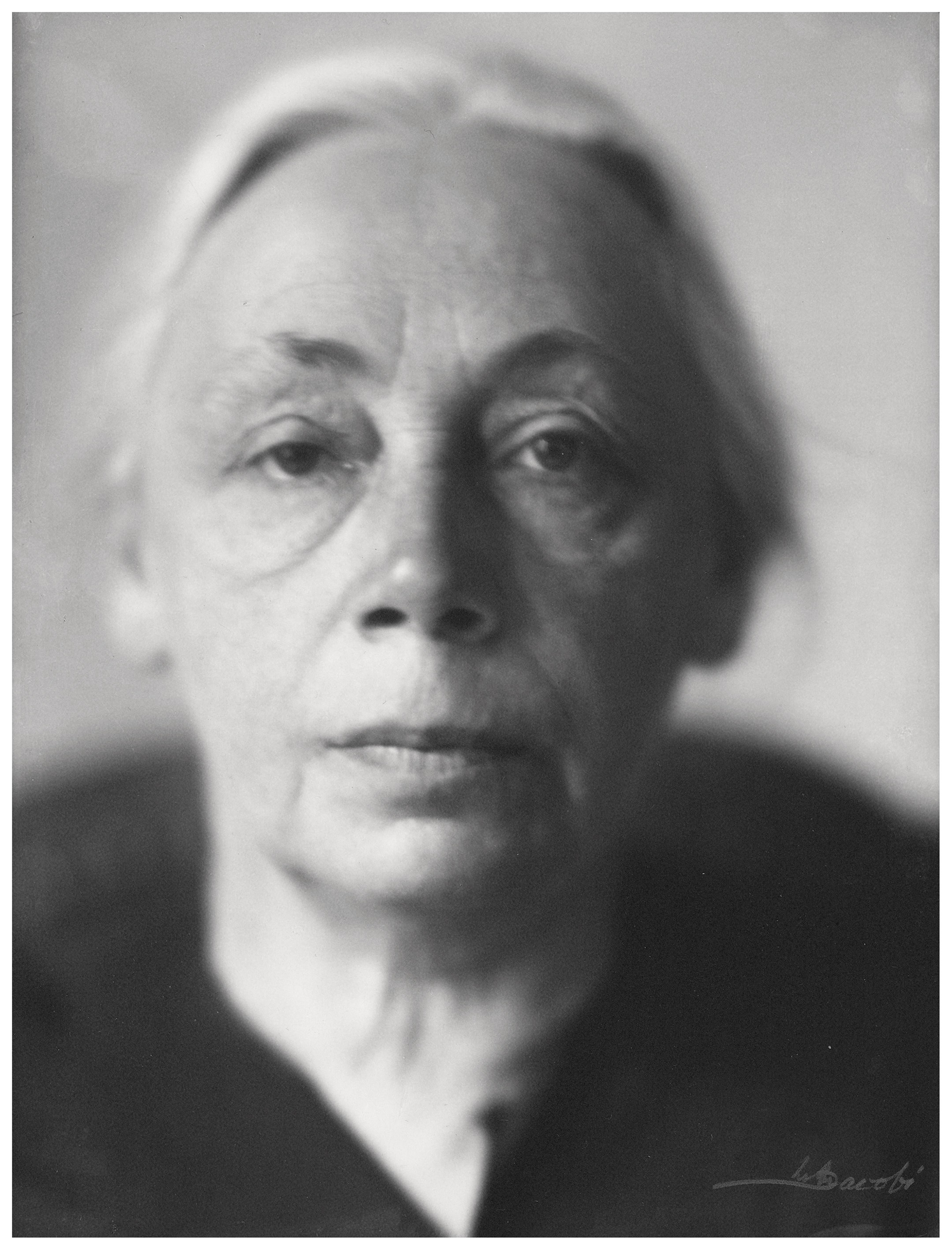 This photograph by Lotte Jacobi from 1929 presents a black-and-white portrait of the artist Käthe Kollwitz in close-up, with her face filling almost the entire frame. Her gaze is calm and directed straight at the camera, while her expression conveys a serious, deeply experienced, and dignified presence. Light falls from the side, accentuating the fine lines of her face and creating a strong contrast between her illuminated features and the dark background.