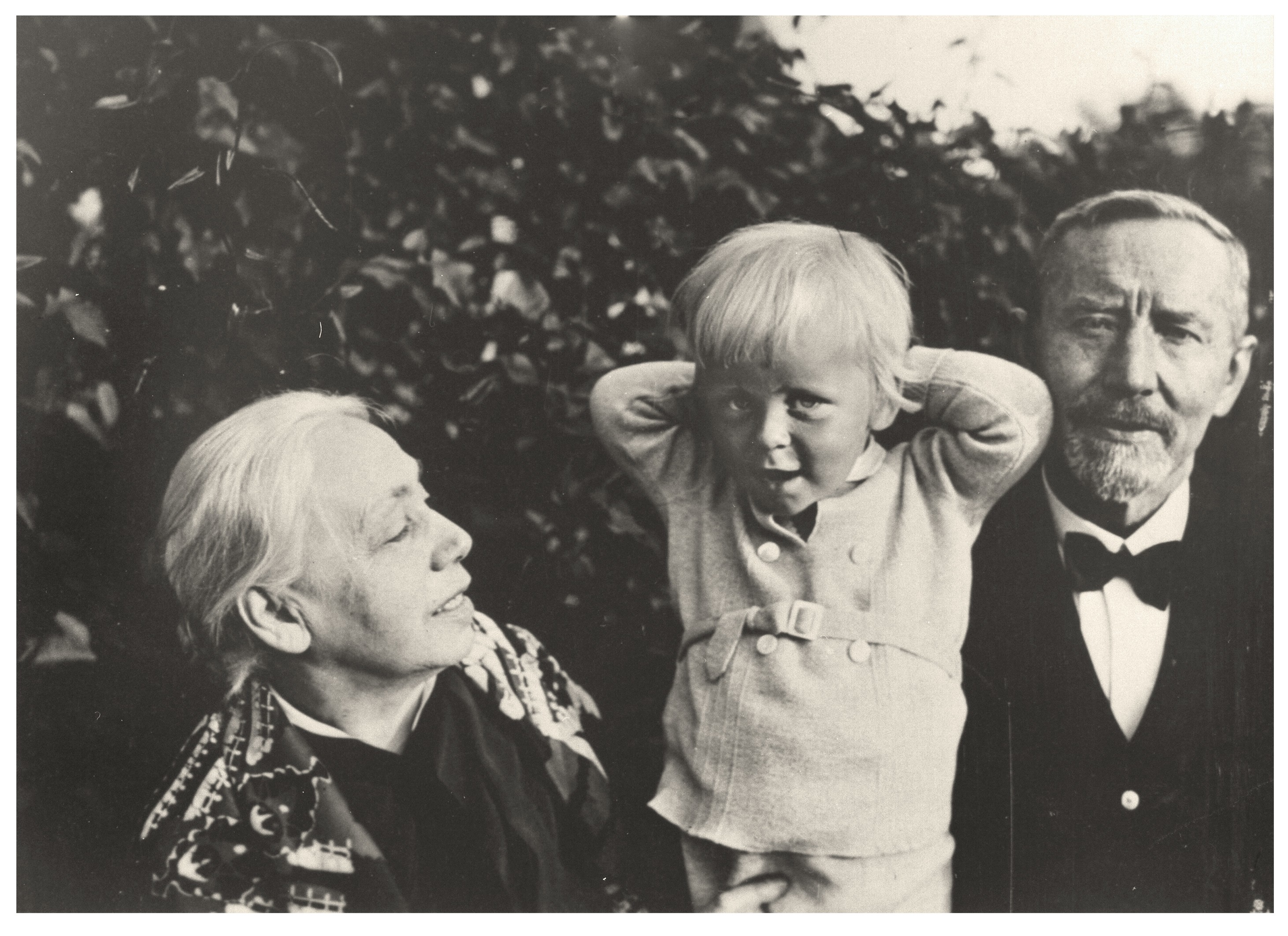 The black-and-white photograph shows Käthe Kollwitz (left), her husband Karl (right), and their grandson Arne (center) in a garden, with dense shrubbery in the background. Käthe Kollwitz looks up at the blond child with a smile as he holds both hands behind his head, while Karl, a bearded man wearing a dark suit and bow tie, stands to the right and looks directly into the camera. The scene conveys a private and intimate family moment captured in a family portrait.