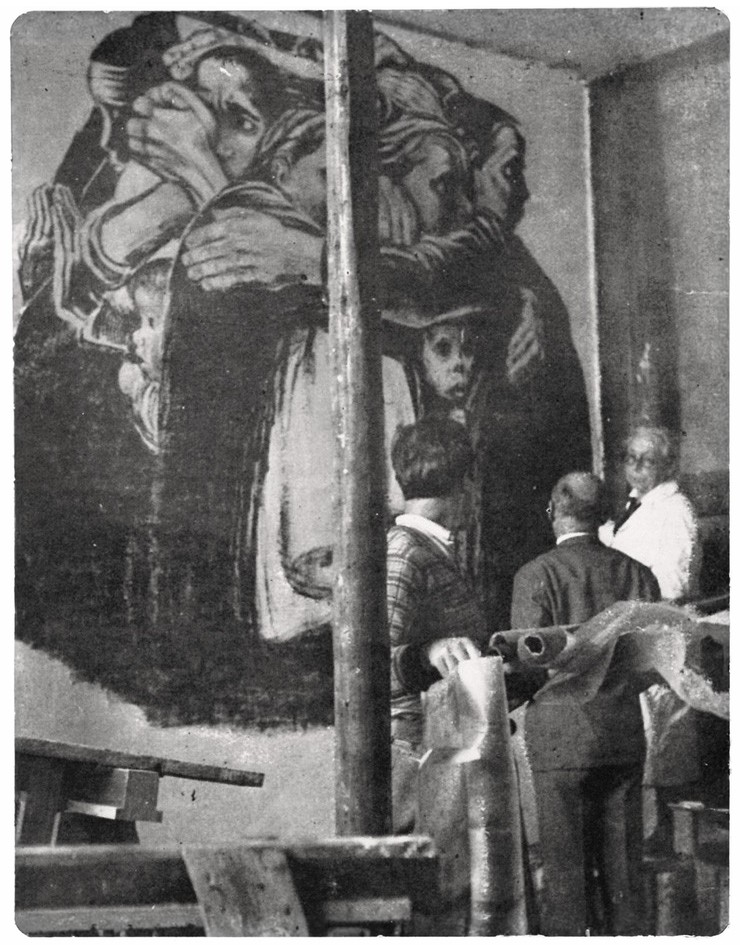 Historic black-and-white photograph showing a large mural on an interior wall depicting a group of women closely embracing one another while protectively holding small children at the center. In the foreground, Käthe Kollwitz stands with two men in period clothing as they observe and work on the monumental artwork. The scene documents the installation process and emphasizes the scale and emotional intensity of the composition.