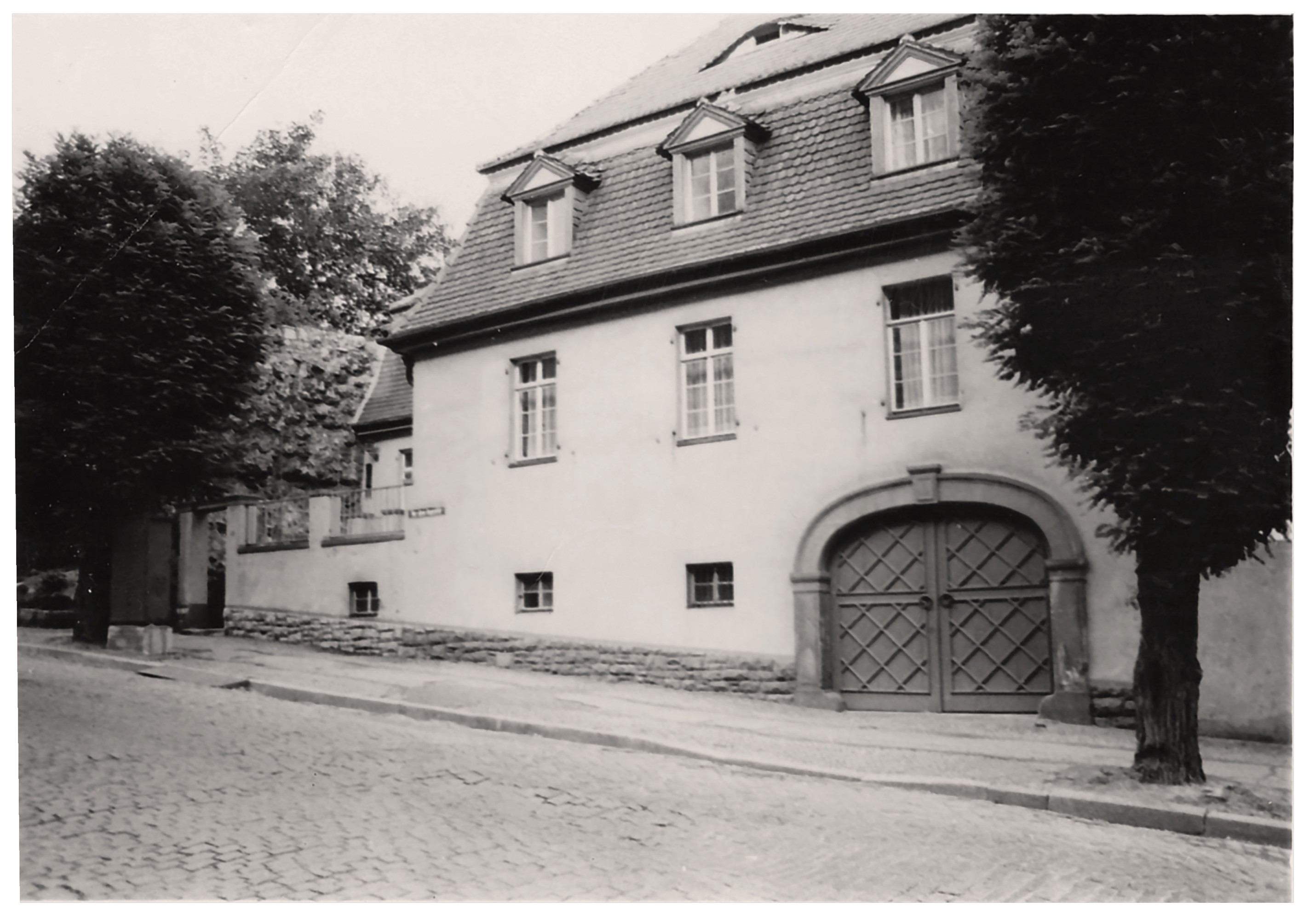 The image shows a light-colored, two-story corner house on a cobblestone street, topped by a dark mansard roof with three small dormer windows. On the ground floor, to the right, there is a large round-arched wooden gate with a diamond pattern, while several simple windows are arranged in the façade to the left. The building is framed on both sides by tall, densely leafed trees, which partially cast the house in shadow.