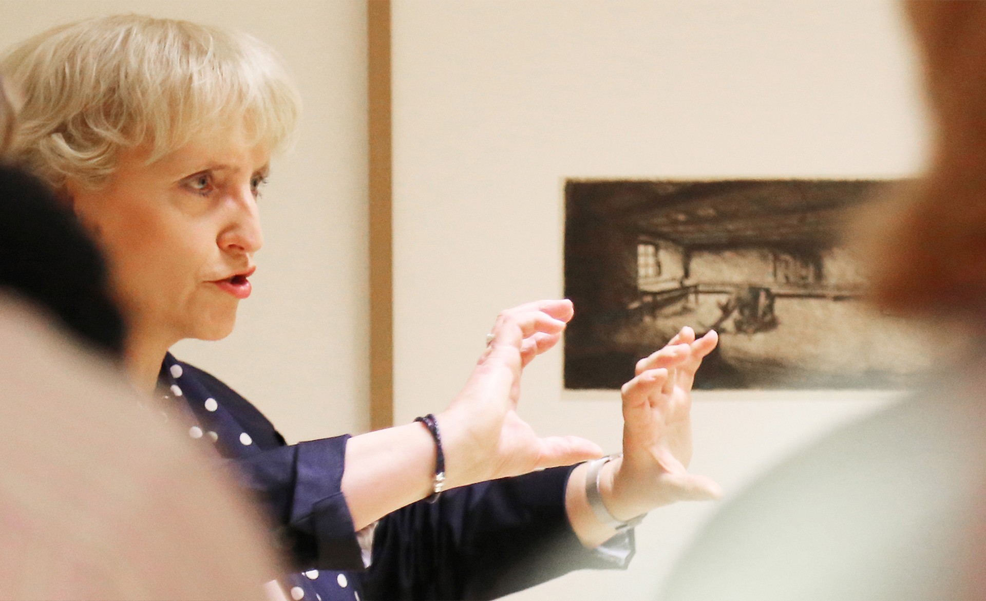 Das Bild zeigt eine Kunstvermittlerin im Profil, die mit lebhafter Mimik und weit geöffneten, erklärenden Händen vor Käthe Kollwitz’ Zyklus „Ein Weberaufstand“ steht. Während sie zu einer Gruppe spricht, blickt man an den verschwommenen Silhouetten einiger Besucherinnen vorbei direkt auf sie und eine dunkle Grafik im Hintergrund.