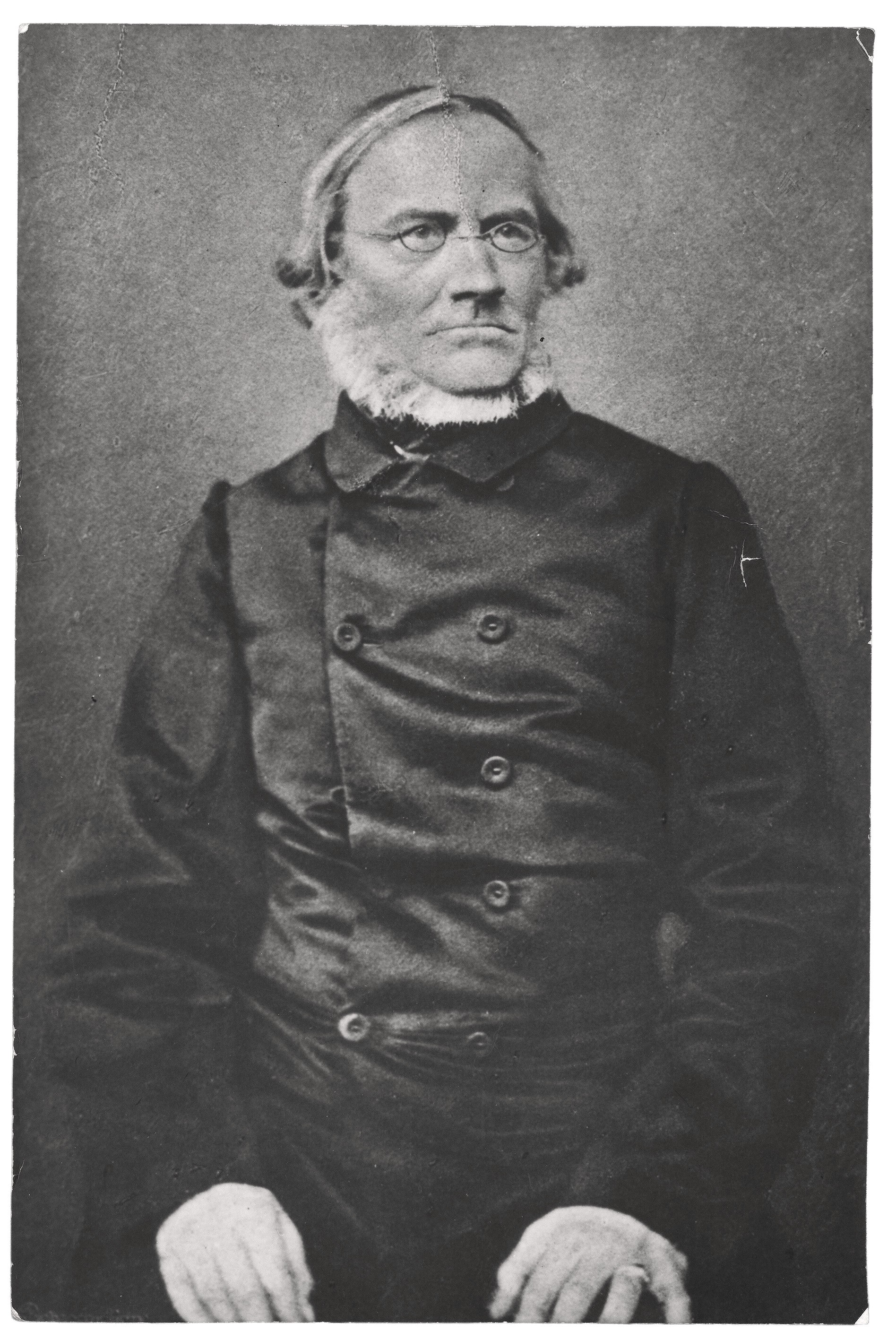 Das Schwarz-Weiß-Porträt zeigt Julius Rupp (1809–1884) als älteren Mann, der mit einem ernsten, leicht zur Seite gewandten Blick dargestellt ist. Er trägt eine dunkle, hochgeschlossene Jacke mit auffälligen Knöpfen, eine schmale Brille und einen markanten weißen Backenbart, der sein Gesicht unterhalb des Kinns wie einen Rahmen umschließt. Seine Hände ruhen ruhig im unteren Bildvordergrund auf seinen Knien.