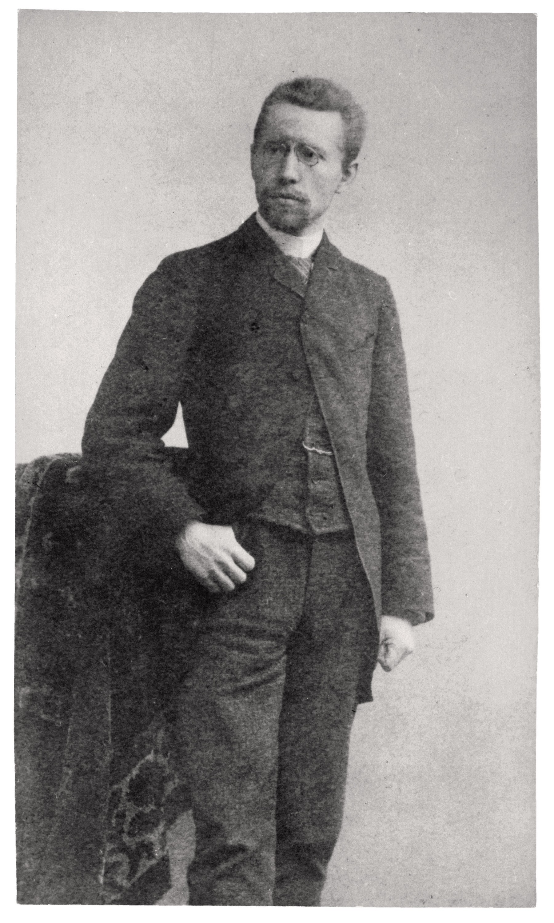 Das Bild zeigt ein historisches Schwarz-Weiß-Porträt von Karl Kollwitz, um 1885, als schlanken Mannes in stehender Pose. Er trägt einen dunklen Gehrock über einer Weste, hat einen kurzen Vollbart sowie eine Brille und stützt seine rechte Hand lässig in die Hüfte. Sein Blick ist ernst und leicht am Betrachter vorbei in die Ferne gerichtet, während er vor einem neutralen, hellen Hintergrund steht.
