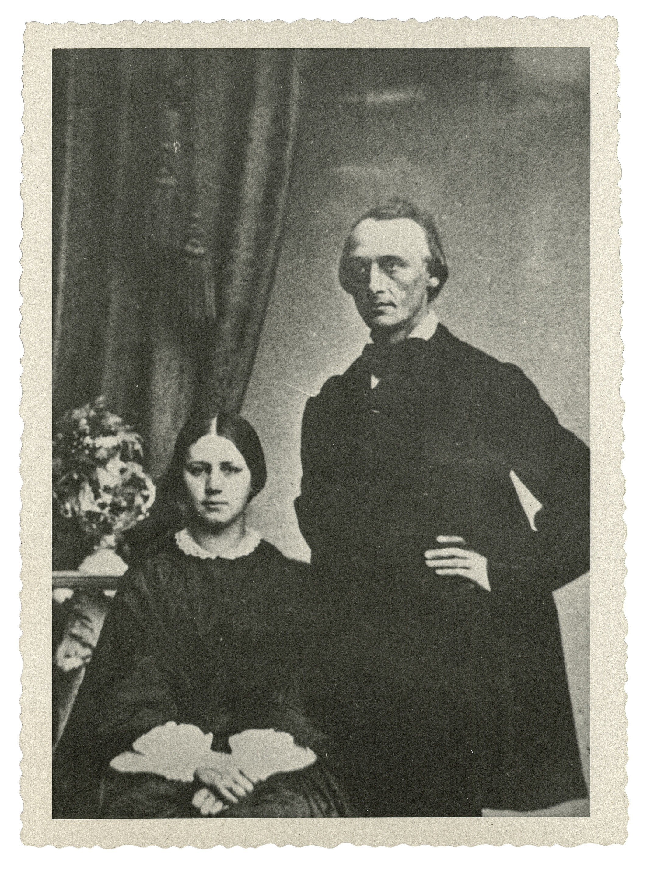 Das Bild zeigt ein Schwarz-Weiß-Porträt von Carl Schmidt (1825–1898), stehend, und seiner Frau Katharina Schmidt, geb. Rupp (1837–1925), links von ihm sitzend, in formeller Kleidung aus der Mitte des 19. Jahrhunderts. Carl Schmidt trägt einen dunklen Gehrock und hat eine Hand in die Hüfte gestützt, während seine Frau in einem dunklen Kleid mit hellem Spitzenkragen und Manschetten ihre Hände im Schoß gefaltet hat. Im Hintergrund sind ein schwerer Vorhang mit Quaste sowie ein kleiner Tisch mit einem Blumenarrangement zu erkennen.