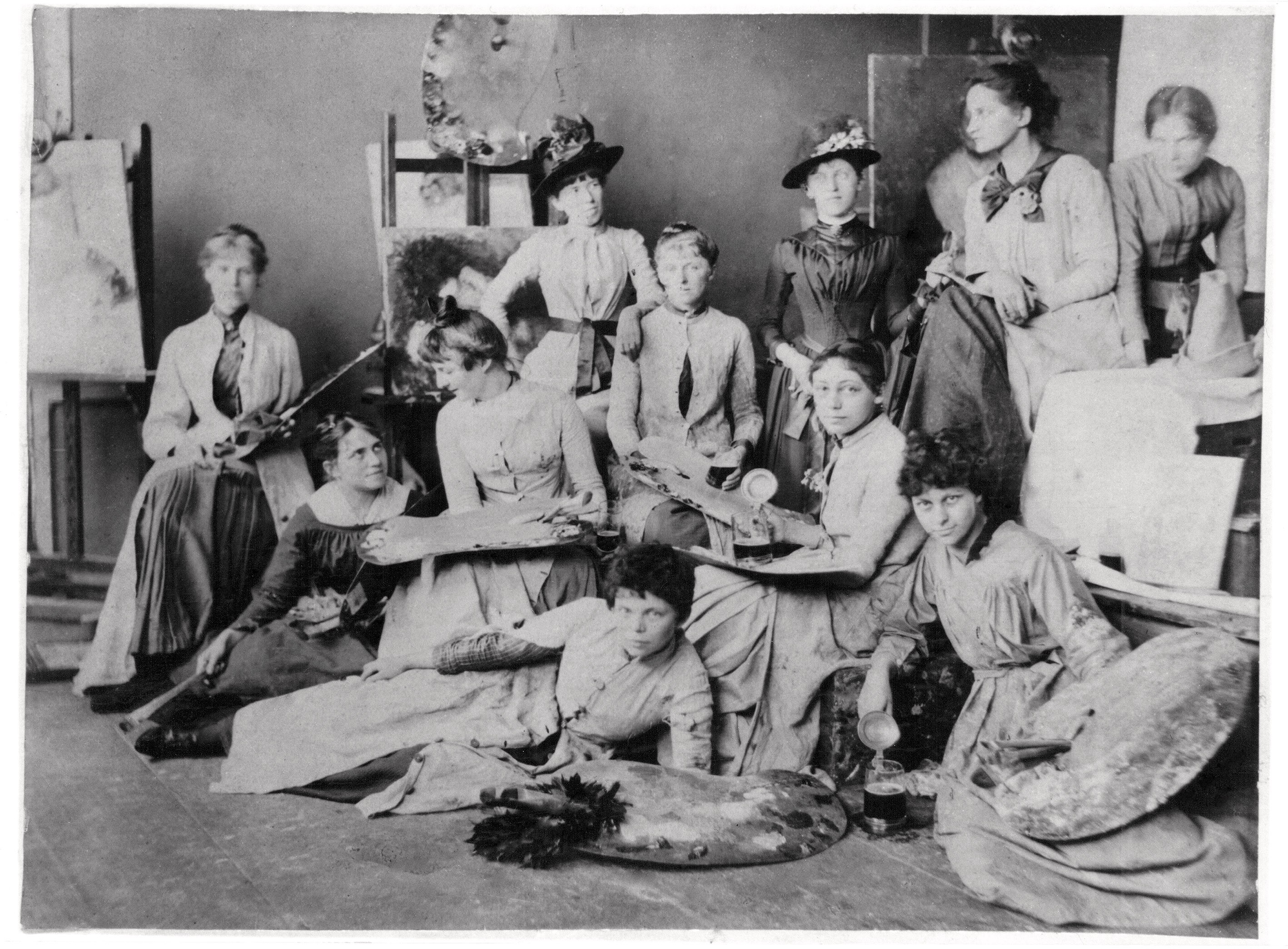 Das Schwarz-Weiß-Foto aus dem Jahr 1889 zeigt eine Gruppe von elf jungen Frauen aus der Münchner Malklasse von Ludwig Herterich, um 1889, Käthe Schmidt sitzend als zweite von rechts, die in lockeren Posen zwischen Staffeleien und Leinwänden im Atelier verteilt sind. Die Frauen tragen hochgeschlossene, zeitgenössische Kleider und halten teilweise Malutensilien wie Paletten oder – als humorvolles Detail – einen Bierkrug in den Händen. Der Raum wirkt lebendig und unkonventionell, wobei einige der Künstlerinnen auf dem Boden sitzen, während andere im Hintergrund stehen oder auf Hockern thronen.