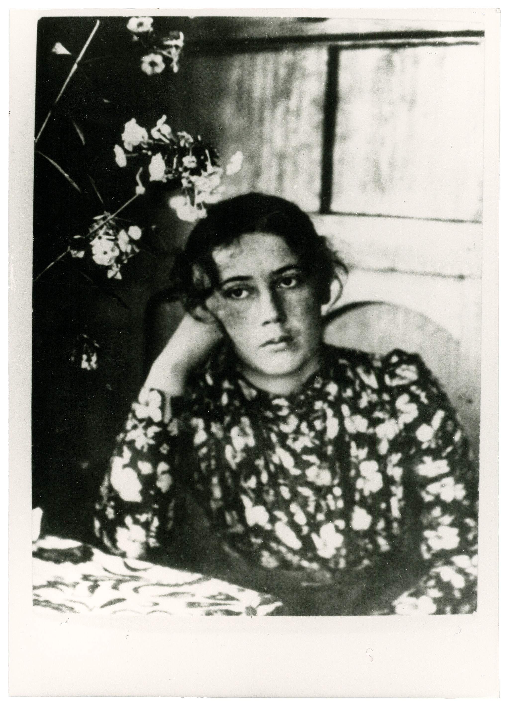 Dieses Schwarz-Weiß-Foto zeigt Lisbeth Stern, geb. Schmidt (1870–1963), die jüngere Schwester von Käthe Kollwitz als eine junge Frau mit dunklem, leicht unordentlichem Haar, die nachdenklich an einem Tisch im Freien, vermutlich auf einer Terrasse, sitzt. Sie trägt ein dunkles Kleid mit einem hellen, kleinteiligen Blumenmuster und stützt ihren Kopf mit melancholischem Blick auf ihre rechte Hand. Links im Vordergrund ragen helle Blüten in das Bild, während der Hintergrund durch ein schlichtes Fenster und eine strukturierte Wand angedeutet wird.