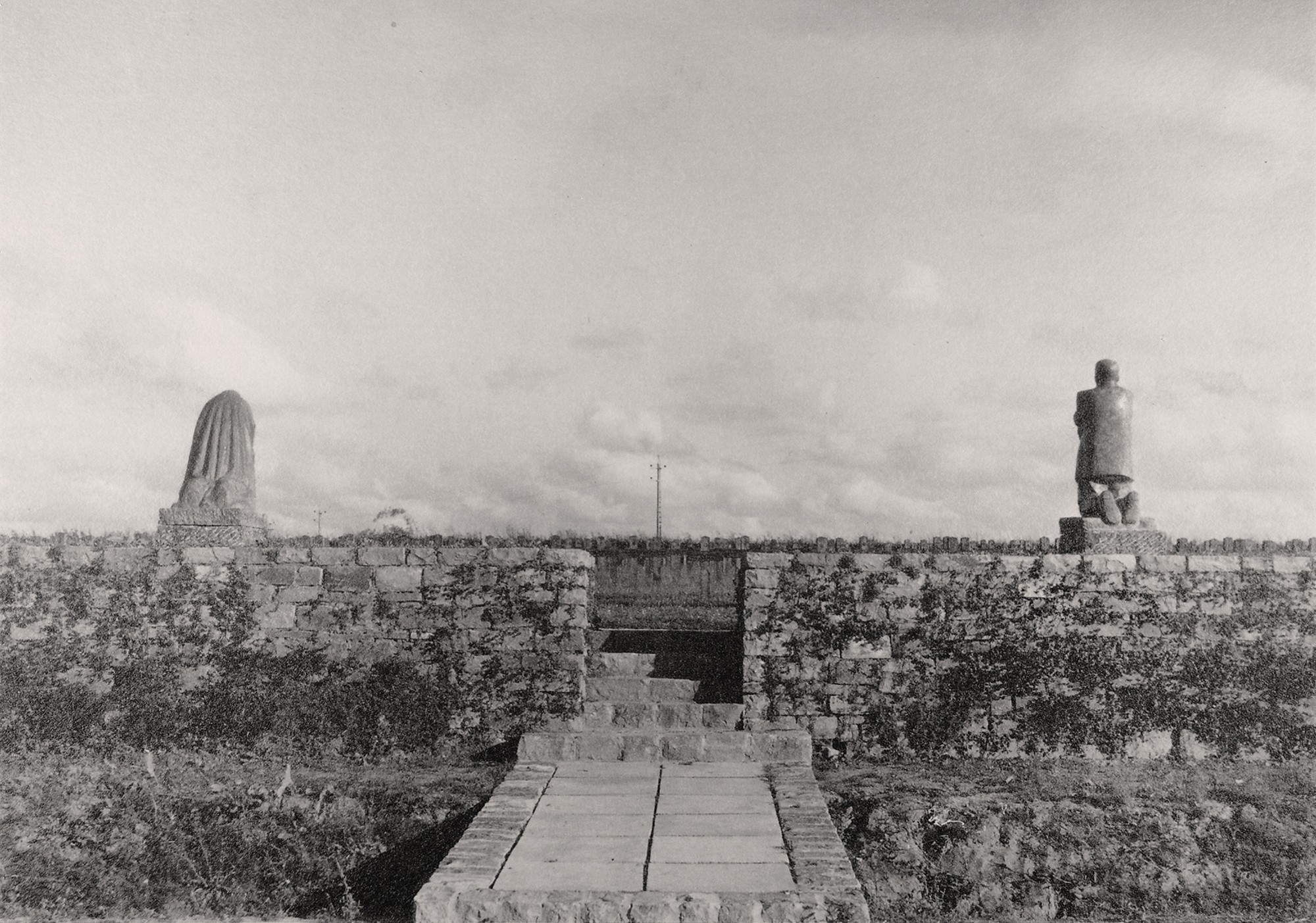 Die Schwarz-Weiß-Fotografie von 1932 zeigt den Blick von Außen auf den Soldatenfriedhof Roggeveld: Ein mit Steinplatten ausgelegter schmaler Weg führt durch eine Öffnung in der niedrigen Bruchsteinmauer in den Friedhof, dahinter rechts vom Eingang die Skulptur der Mutter, links die des Vater, beide von hinten.