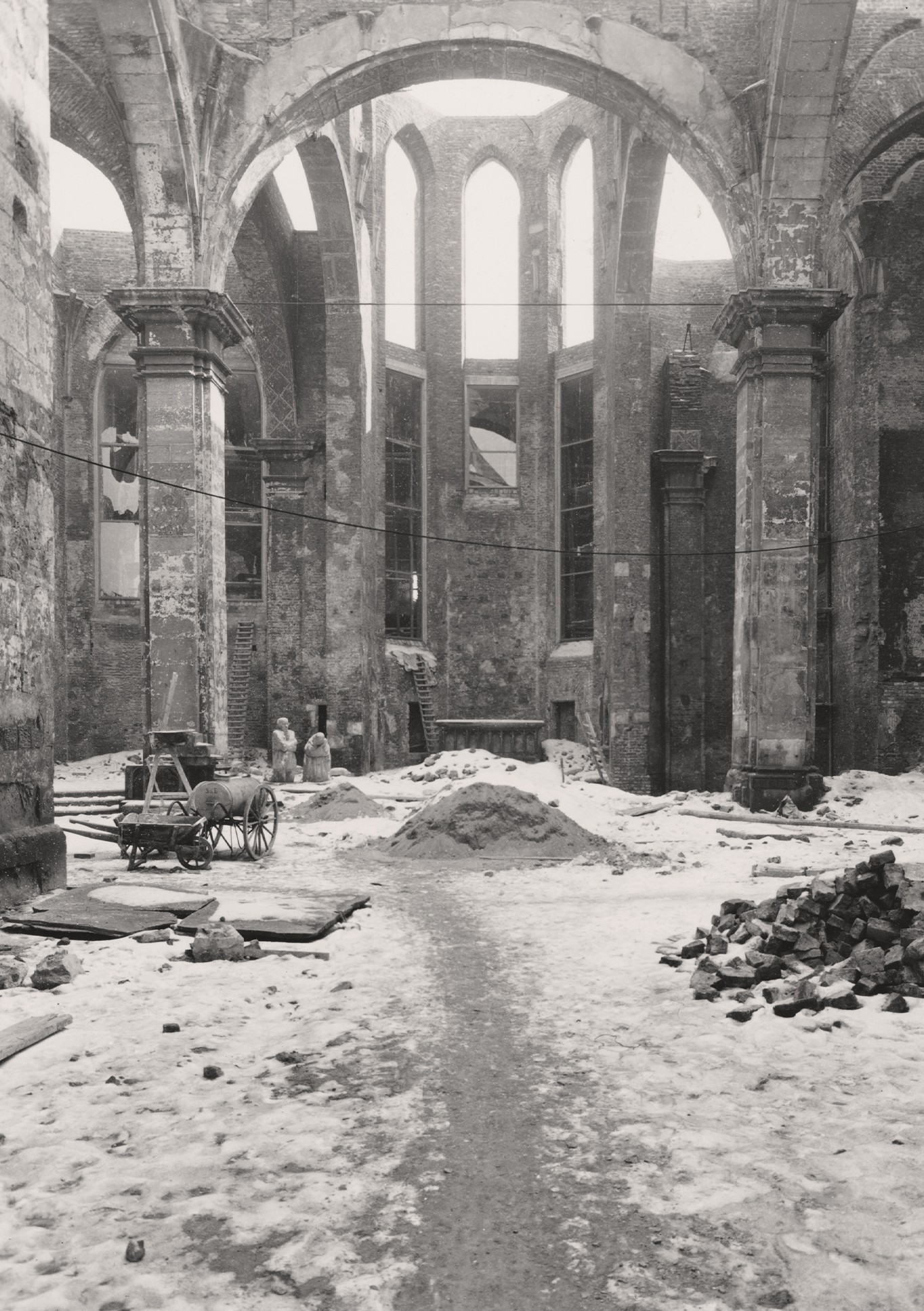 Die Schwarz-Weiß-Fotografie zeigt einen Blick in die Kölner Kirchenruine von Alt St. Alban bei der Aufstellung des Skulpturenpaares »Trauernden Eltern« in den Wintermonaten 1955. Der Innenhof ist schneebedeckt, rechts im Vordergrund ein Steinhaufen, links ein Karren mit großen hölzernen Wagenrädern und Handwerksgerät, im Hintergrund sind die bereits aufgestellten Skulpturen zu sehen.