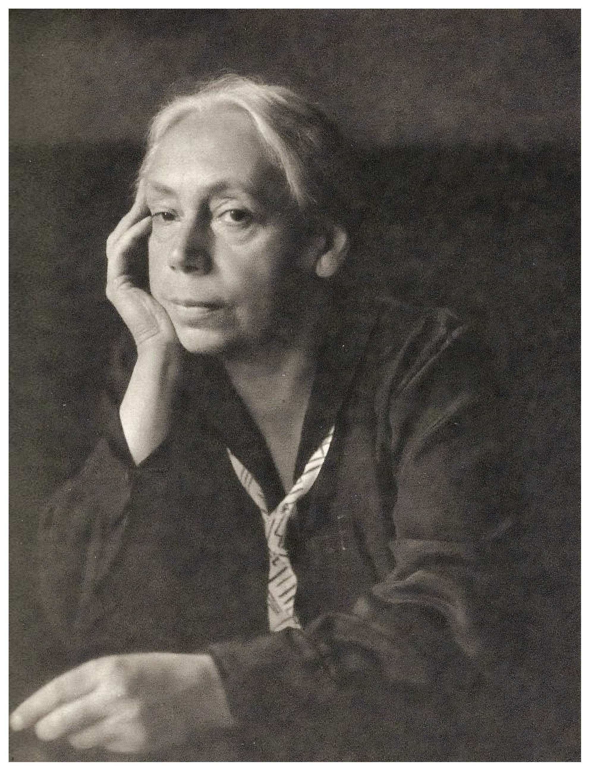 The black-and-white portrait shows Käthe Kollwitz with light-coloured hair combed back, in a pensive pose. She gazes slightly past the viewer with a serious, melancholic expression, whilst resting her head on her right hand. She is dressed in a dark blouse and a light-coloured, patterned bow tie against a calm, dark background.