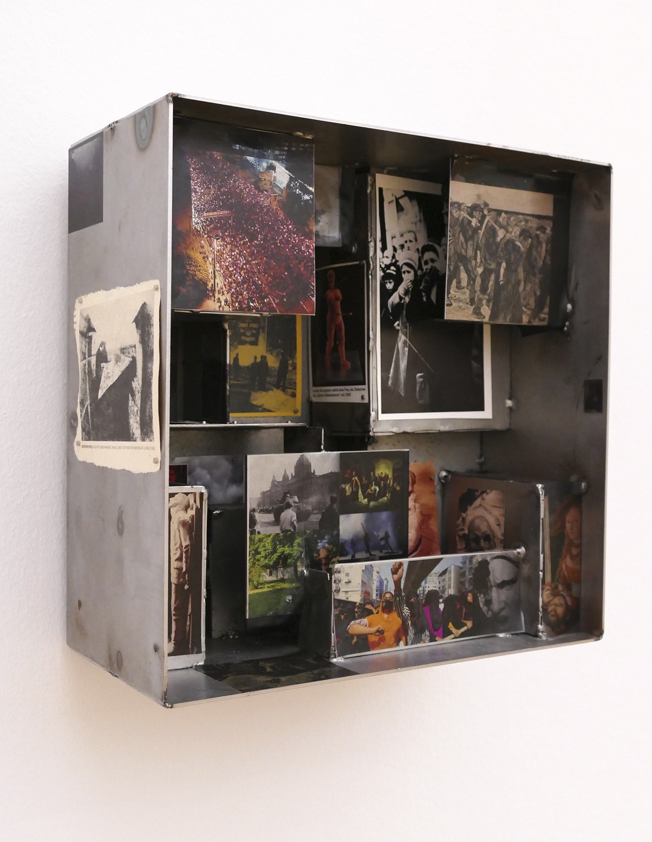 This artwork is a square, open box made of matt grey metal, hanging on a white wall. Inside and on the outer walls, numerous photographs, newspaper clippings and image fragments are arranged in an interlocking pattern, as if in a three-dimensional typesetting case. The images primarily depict mass gatherings, political demonstrations and historical scenes, superimposed on one another across different planes.