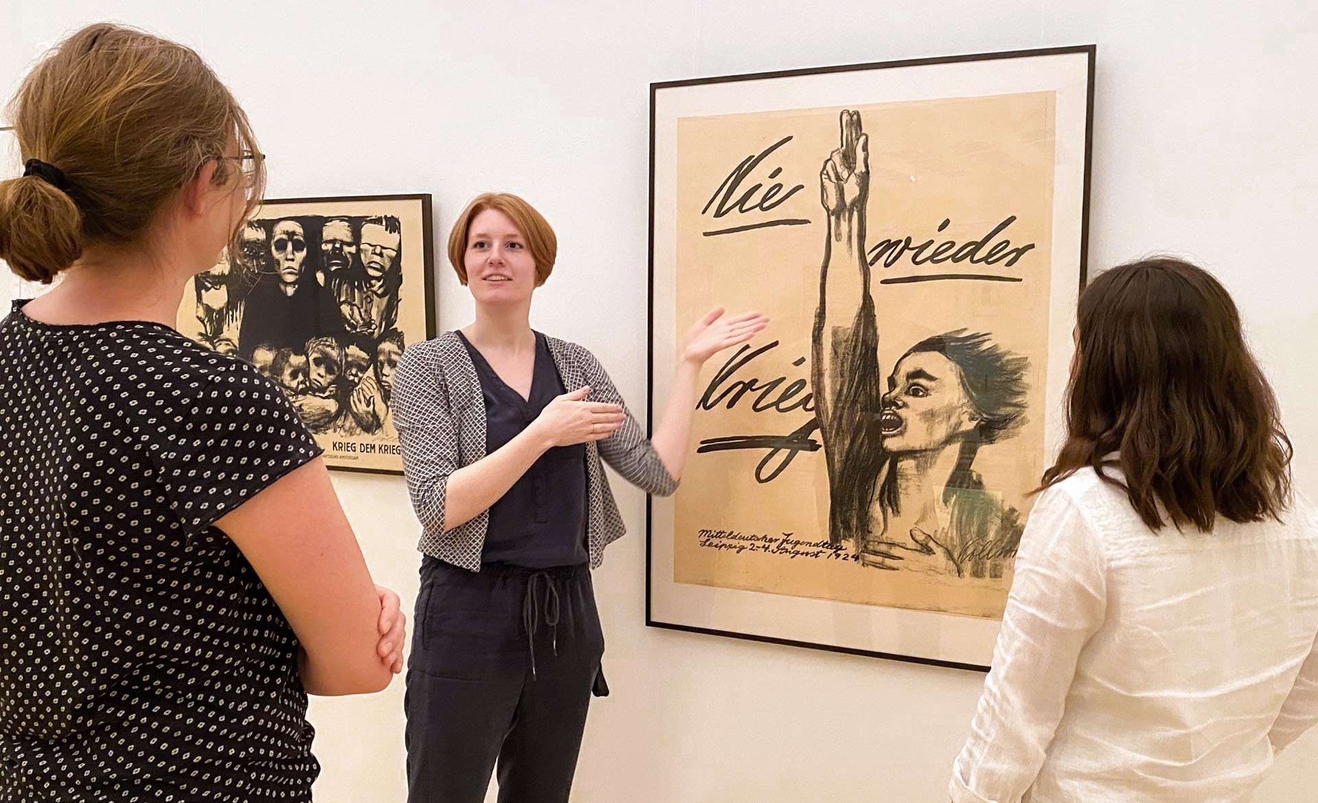 In hellen Ausstellungsräumen des Kollwitz Museum Köln erklärt eine Kunstvermittlerin mit ausladenden Handbewegungen zwei aufmerksam zuhörenden Besucherinnen das berühmte Antikriegsplakat "Nie wieder Krieg" von Käthe Kollwitz. Das Werk steht im Fokus und zeigt die ausdrucksstarke, schwarz-weiße Kohlezeichnung einer Person, die mit weit aufgerissenem Mund und einer zum Schwur erhobenen Hand leidenschaftlich „Nie wieder Krieg“ fordert.