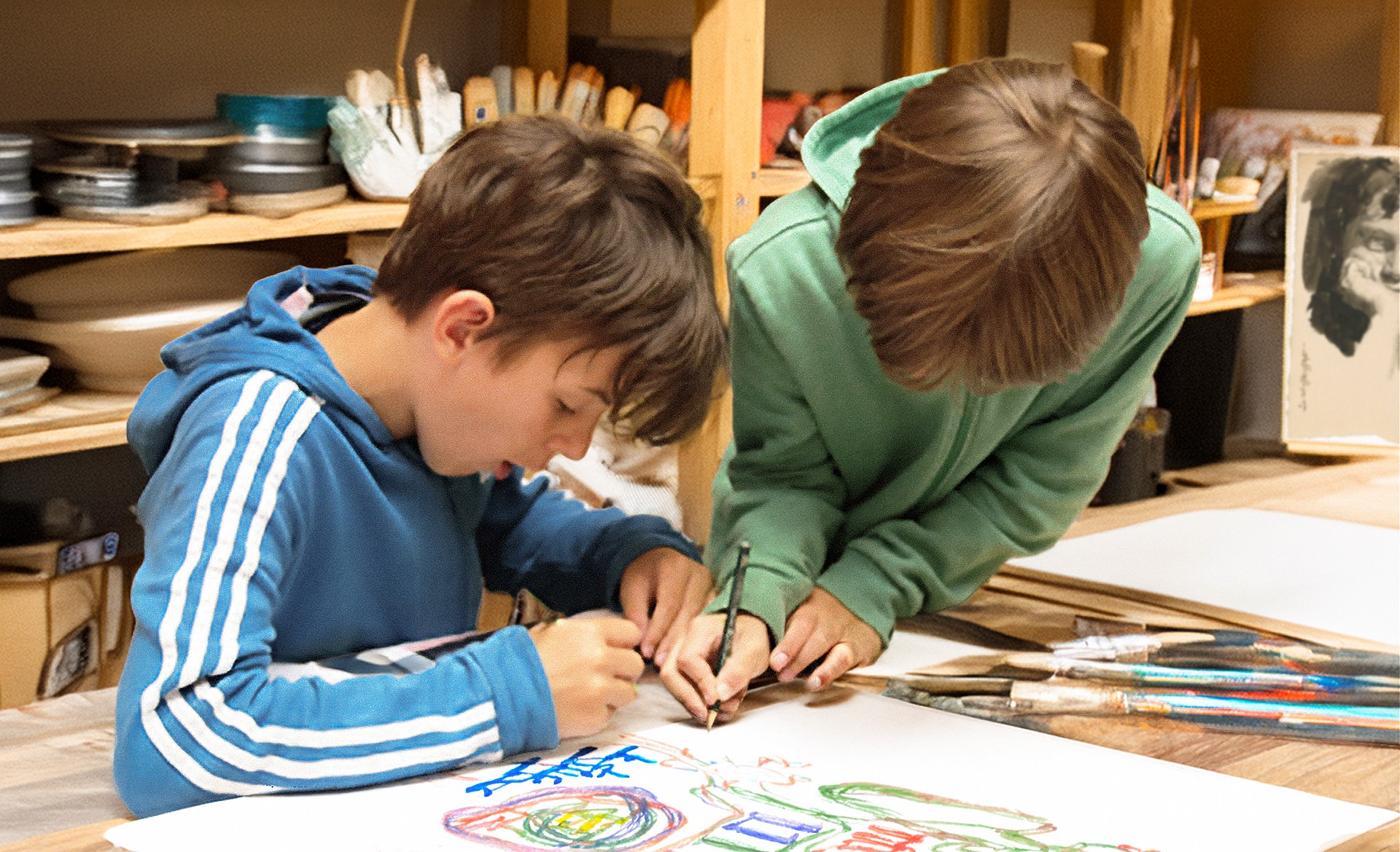 Zwei Jungen beugen sich gemeinsam über einen großen Tisch in der Mitmach-Werkstatt des Kollwitz Museum Köln und zeichnen konzentriert mit bunten Stiften auf ein weißes Blatt Papier. Im Hintergrund sind Regale mit zahlreichen Kunstutensilien wie Pinseln, Farbtöpfen und Skizzen zu sehen, die eine kreative Workshop-Atmosphäre schaffen.