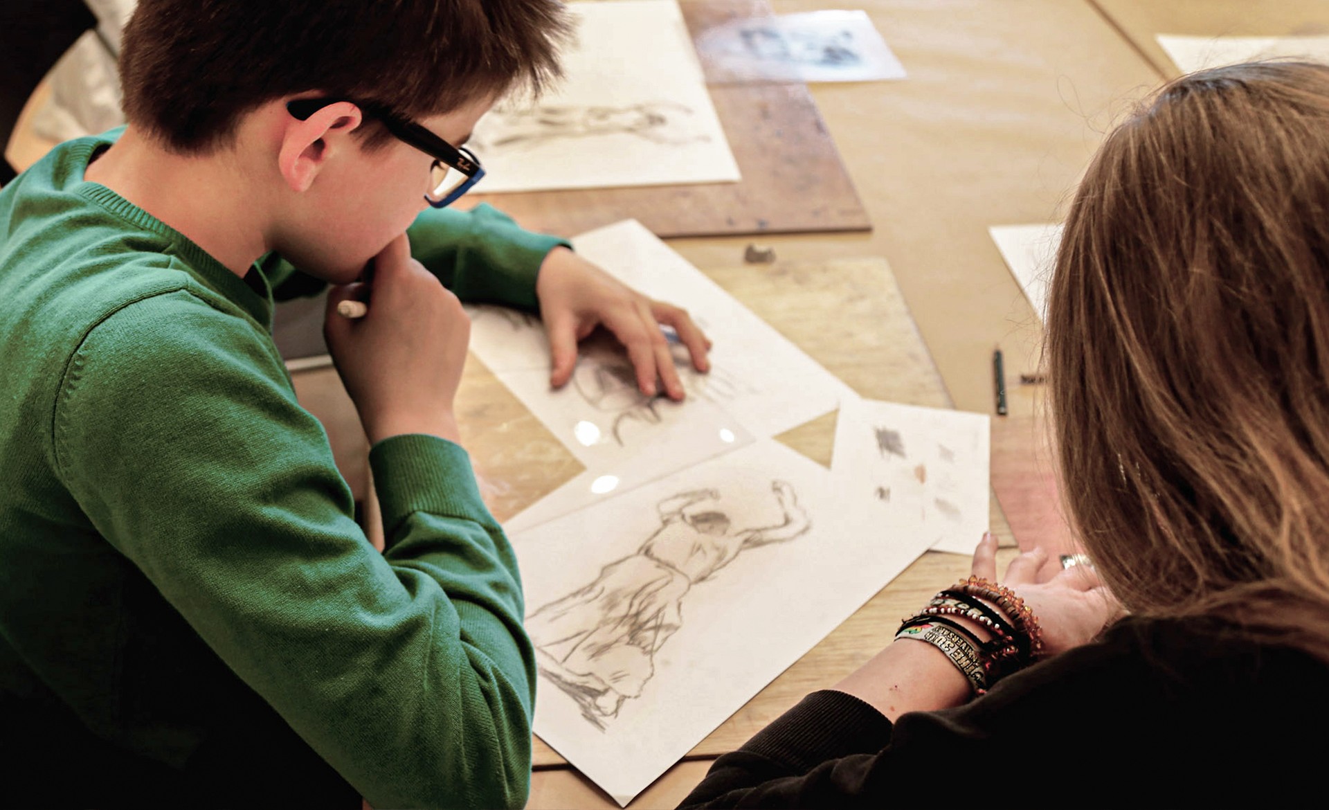 Das Bild zeigt einen Jungen im Profil und die Schulter eines Mädchens, die gemeinsam an einem Tisch über Plexiglasscheiben gebeugt sind. Konzentriert ritzen sie mit feinen Werkzeugen eine Szene aus Käthe Kollwitz' Zyklus „Bauernkrieg“ in das Material, um eine Druckplatte für einen Tiefdruck zu erstellen.